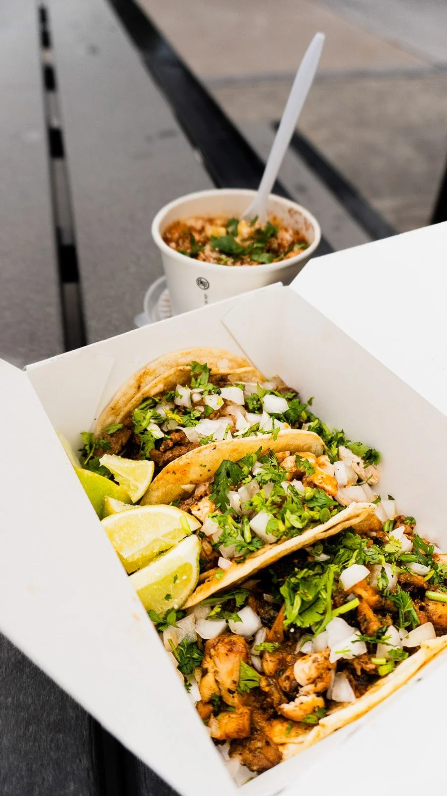 Three tacos in a white takeout box garnished with chopped onions, cilantro, and lemon wedges, with a cup of soup in the background on a black table.