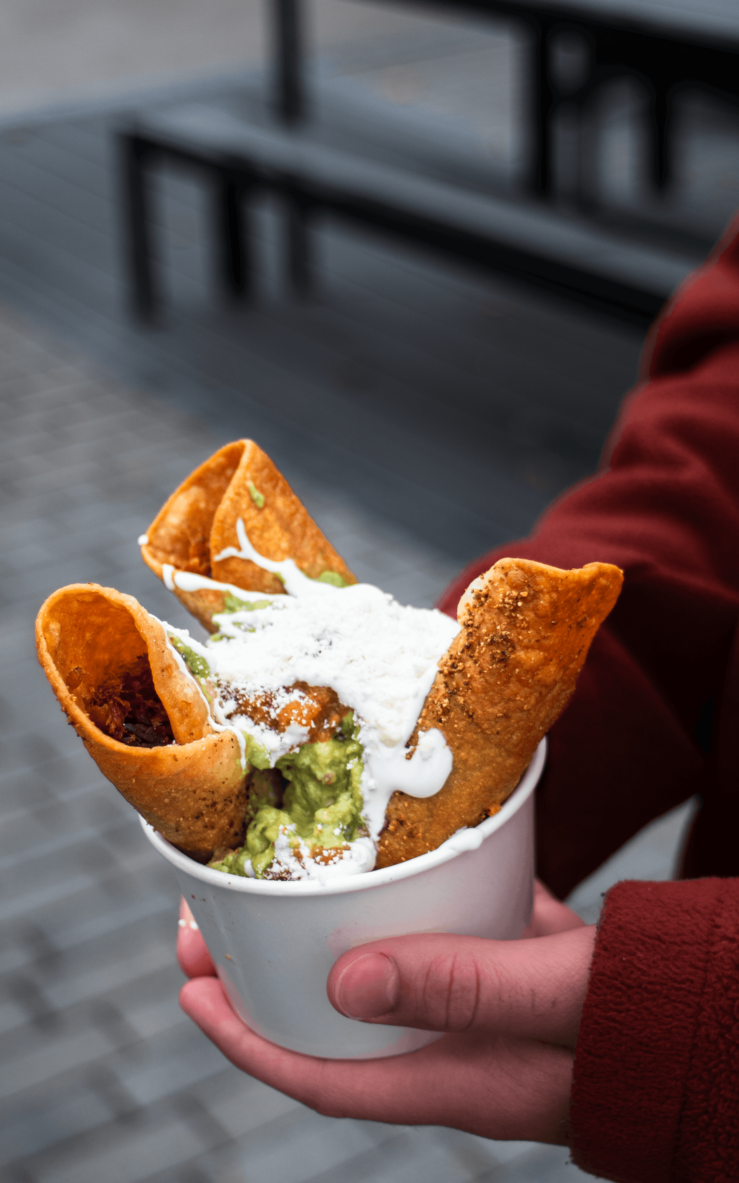 A person holding a white cup filled with Mexican cuisine, including guacamole, sour cream, and four fried tortilla chips, outdoors on a tiled surface with benches in the background.
