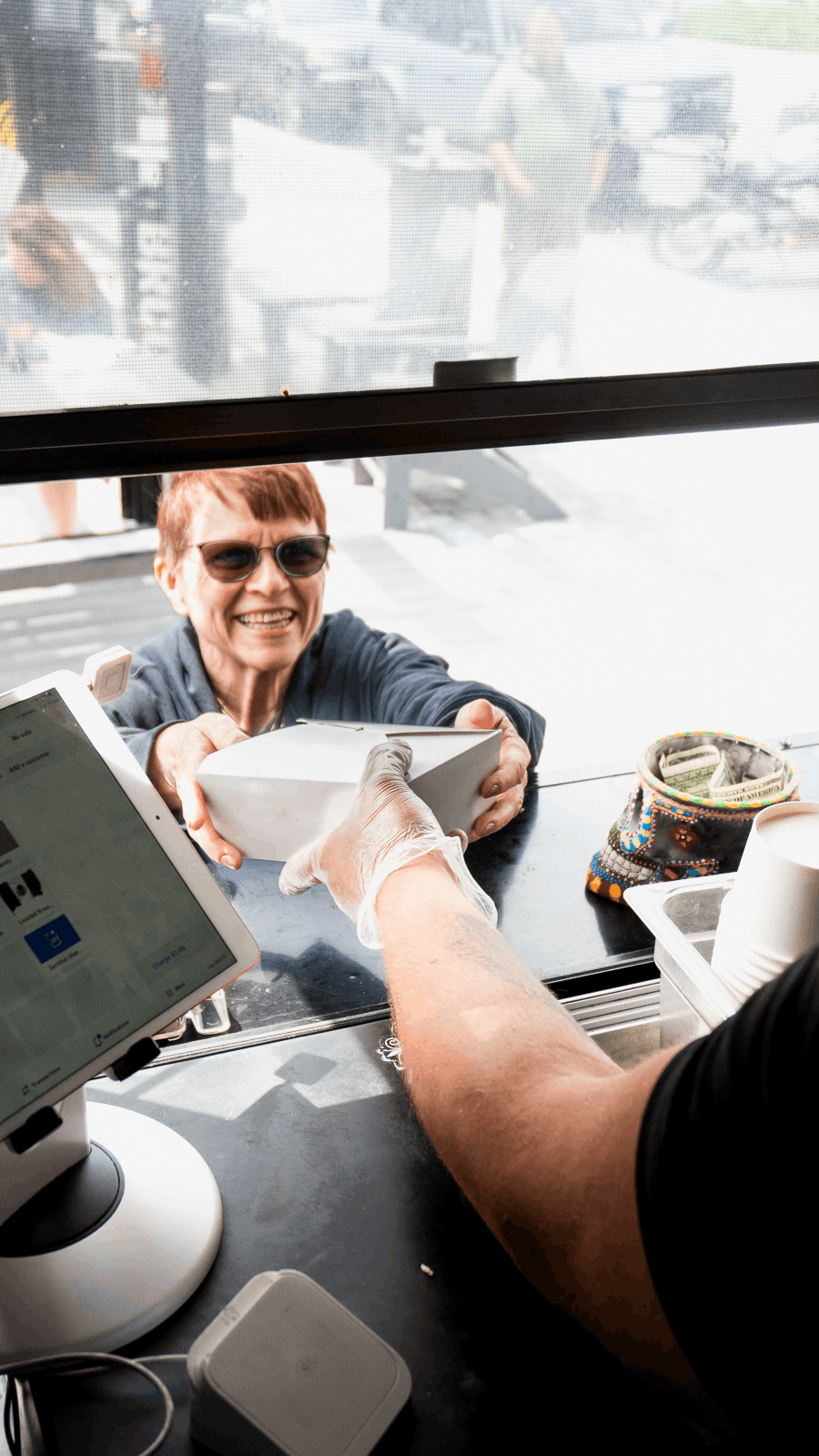 A woman wearing sunglasses is smiling and accepting a paper bag from a person behind the counter at a coffee shop or similar establishment.
