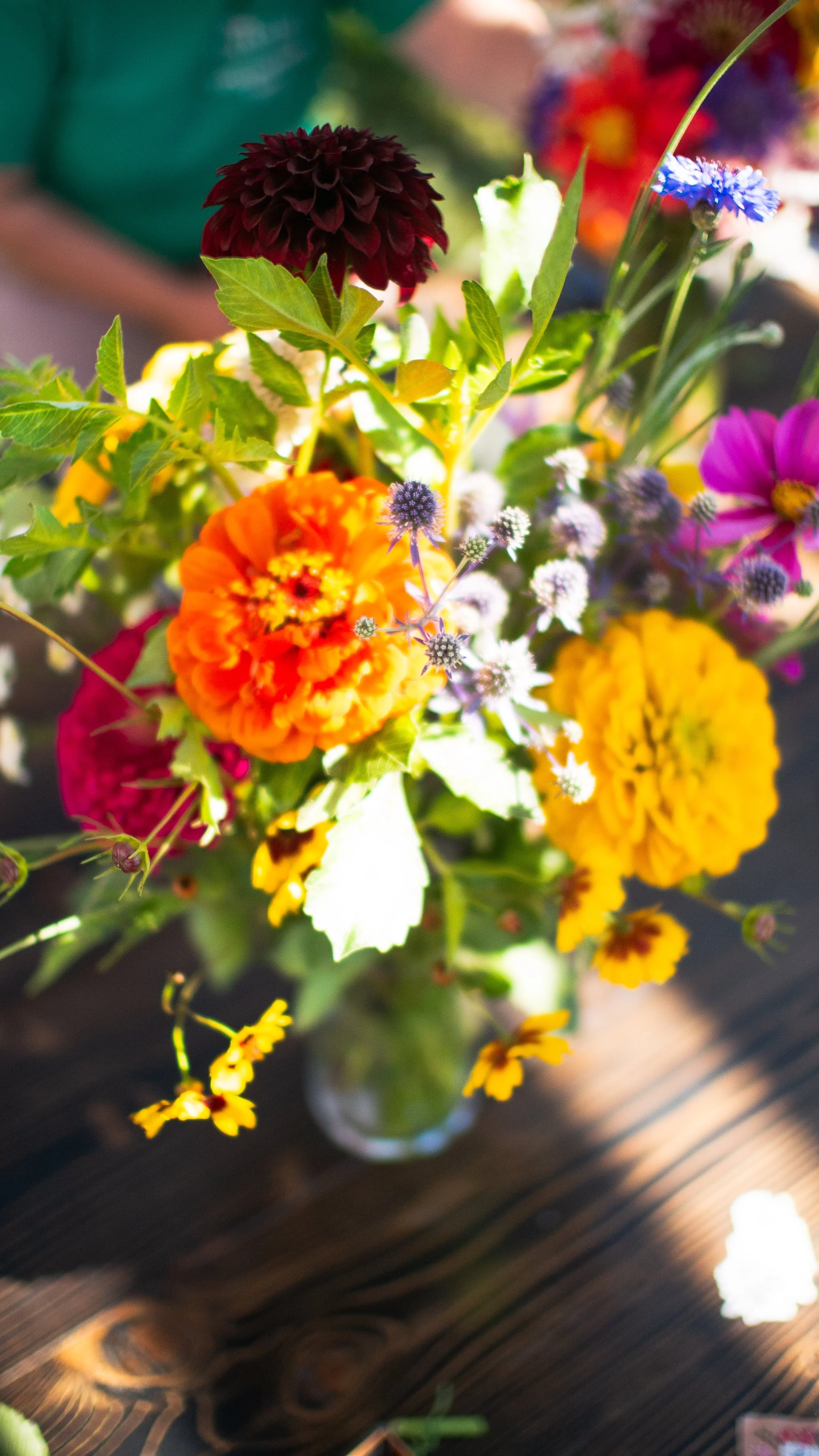A colorful bouquet of various flowers in a glass vase on a wooden surface, with bright and vivid colors including orange, yellow, pink, purple, and burgundy.