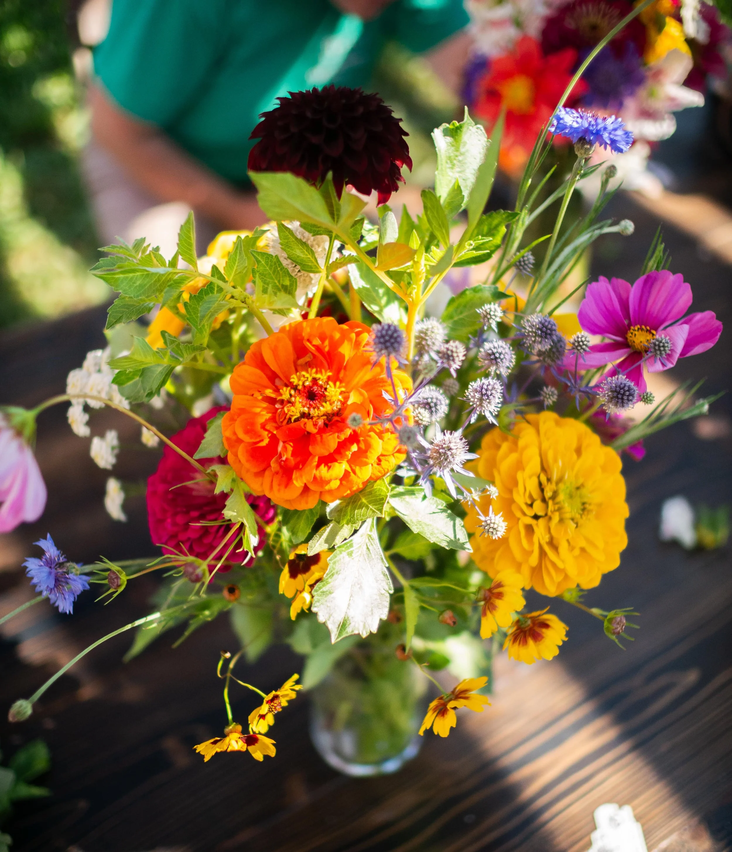 Colorful bouquet of various flowers including marigolds, zinnias, cornflowers, and other wildflowers in a glass vase on a wooden table outdoors.