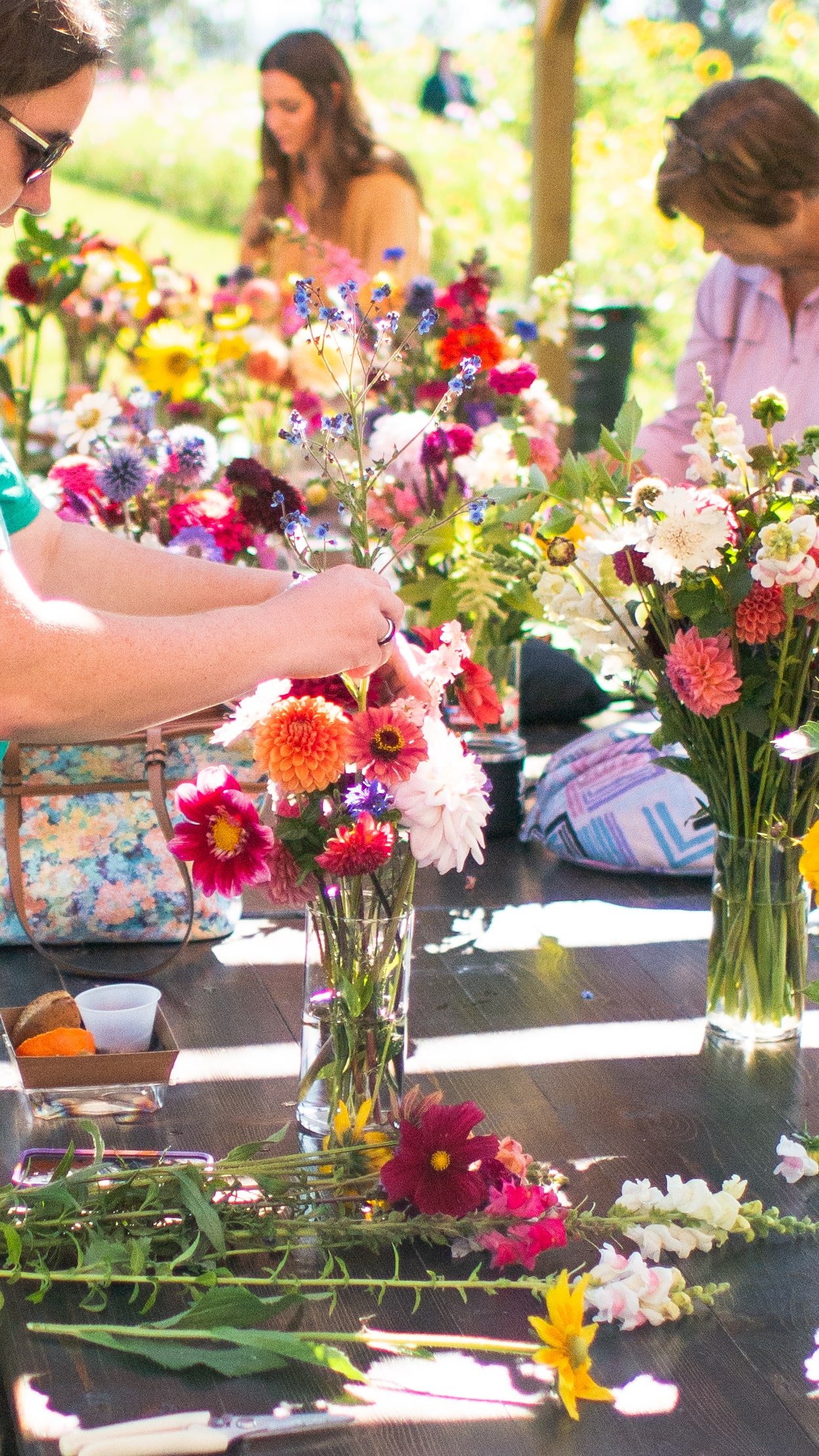 People arranging colorful flowers in glass vases on a wooden table outdoors with sunlight and greenery in the background.