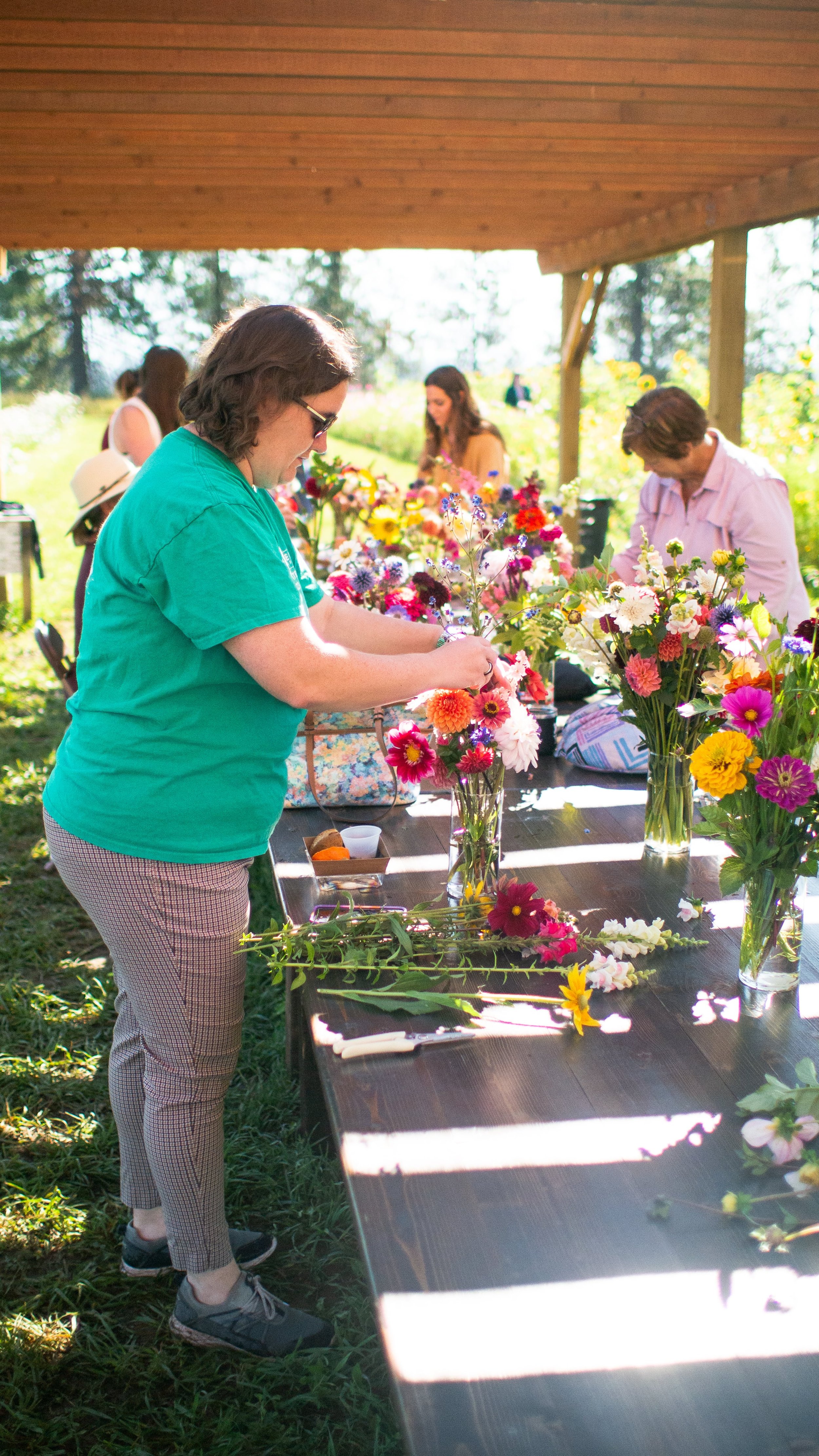 People arranging colorful flowers in glass vases on a long wooden table outdoors, under a wooden roof, with sunlight and trees in the background.