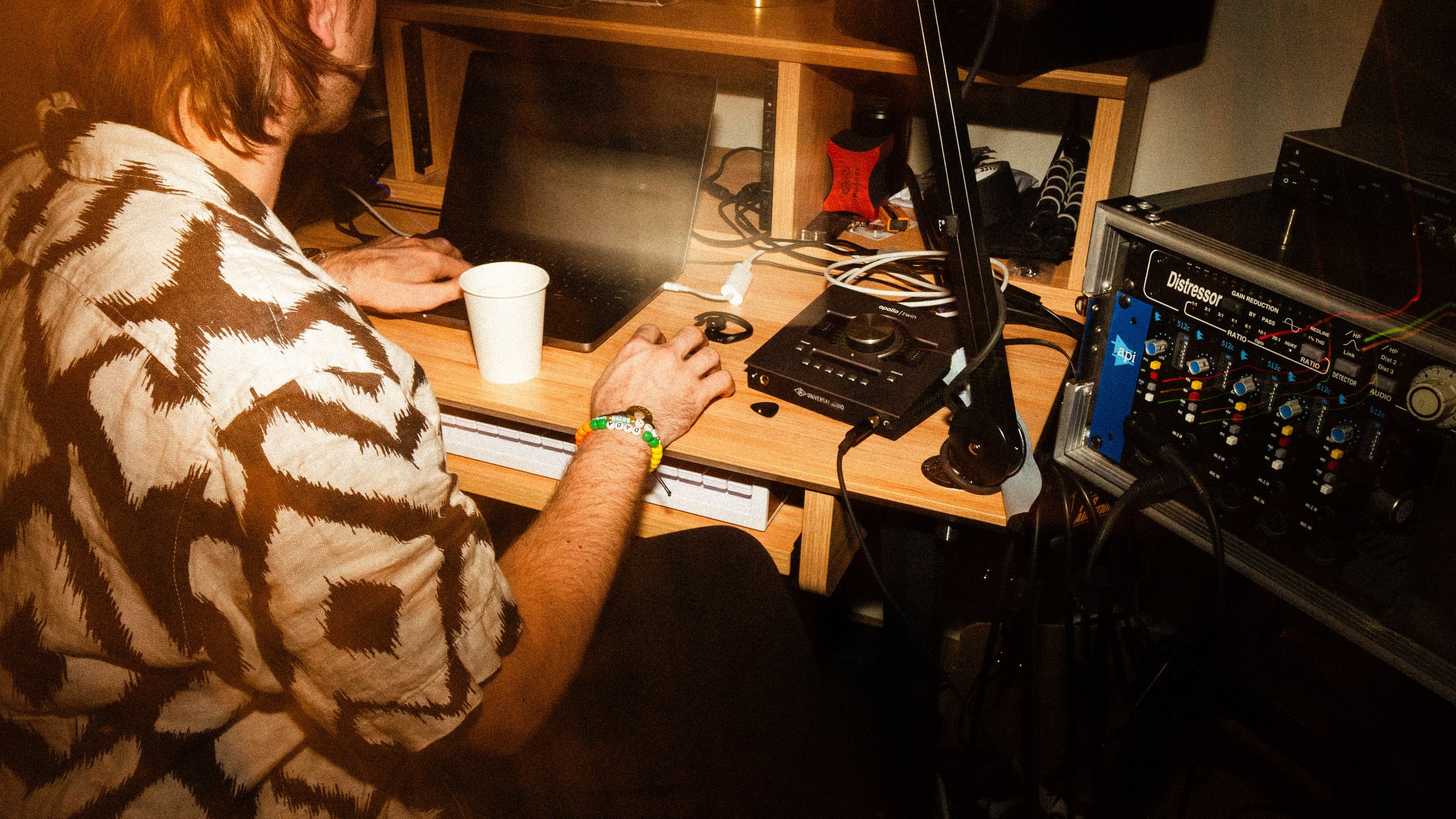 Person with shoulder-length hair wearing a patterned shirt, sitting at a wooden desk, working on a laptop with clubs and electronic music equipment, and a white paper cup nearby.