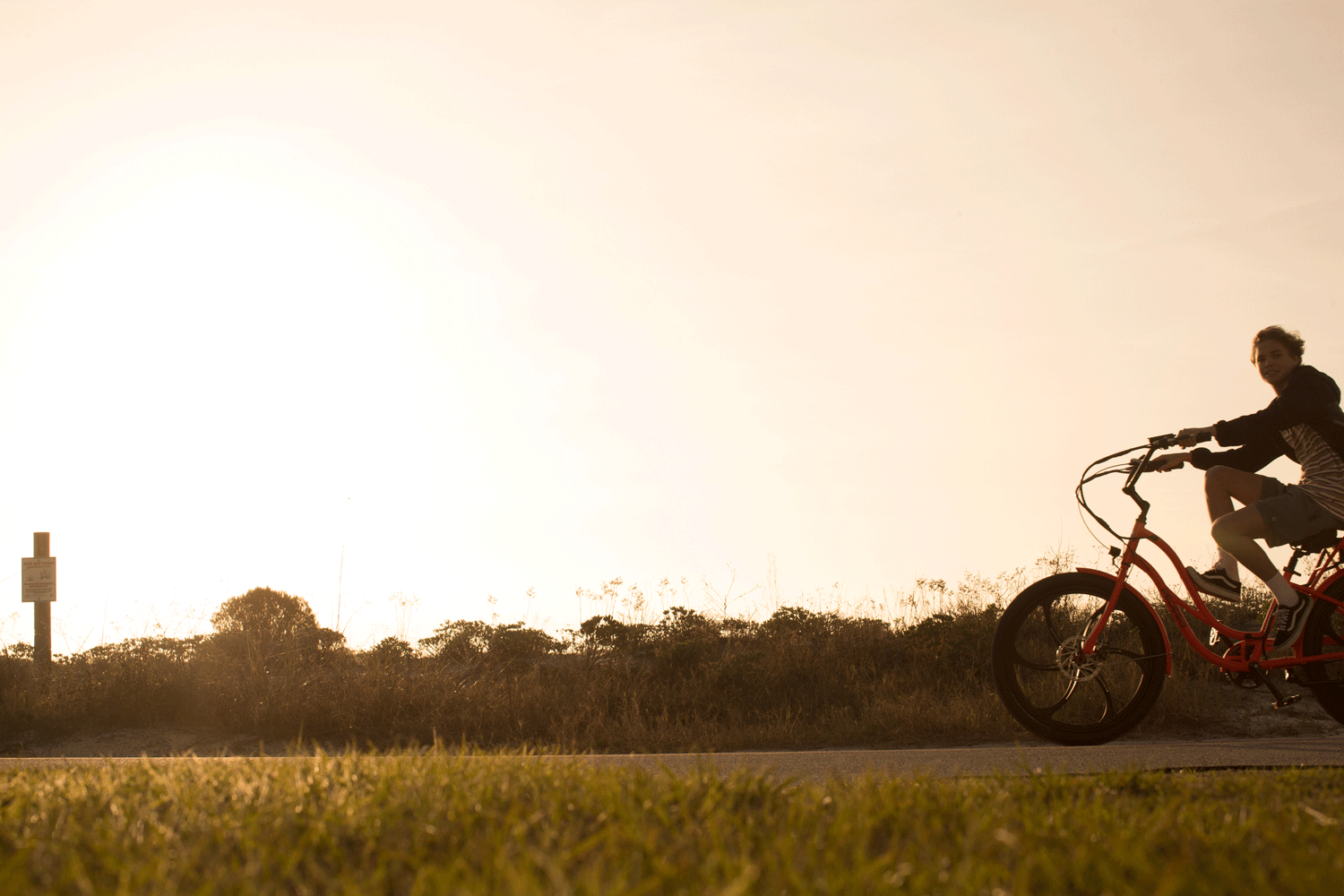 A boy riding a bicycle at sunset on a trail near grassy fields and bushes.