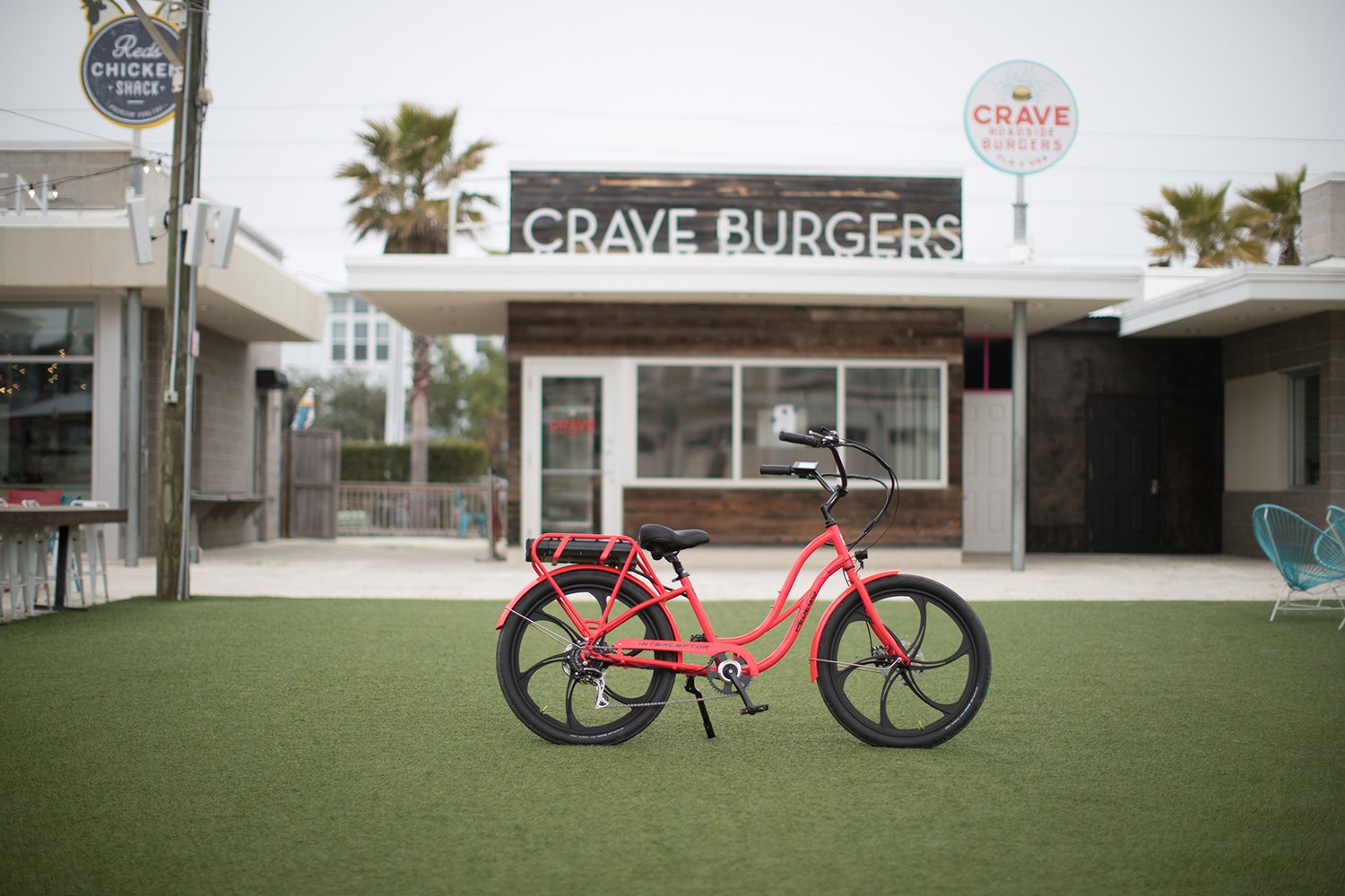 A pink bicycle with a black seat and black wheels is parked on artificial grass in front of a restaurant named Crave Burgers. The restaurant has large windows and a sign on the roof. There is a sign outside that says "Red's Chicken Shack." Palm trees