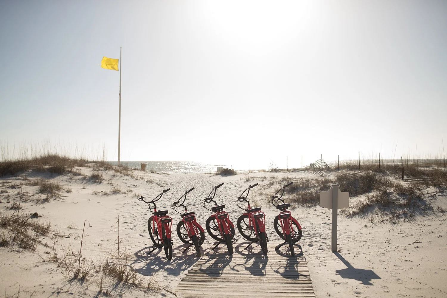 Five red bikes parked on a sandy beach path with a yellow flag flying and sea in the background.