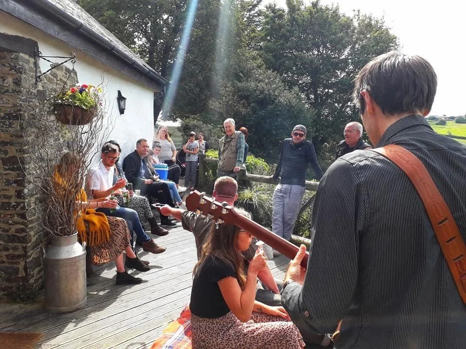 A group of people gathered outdoors on a wooden deck, with some sitting and some standing. A man with a guitar is playing music for a young girl sitting on the deck, who is drinking from a cup. The scene is set near a stone house with plants and tree