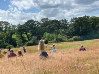 People enjoying a field with tall grass, some sitting and some standing, coasted by trees under a partly cloudy sky.