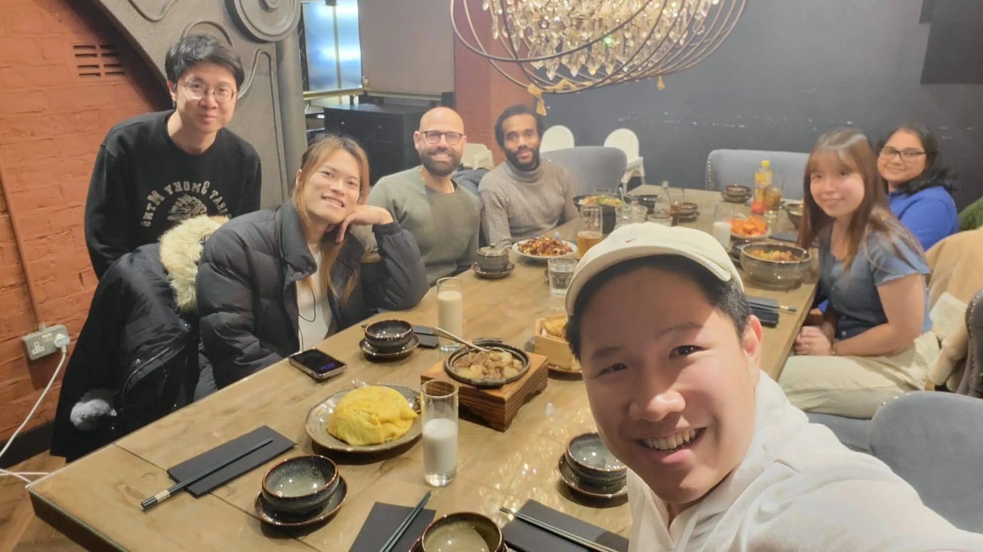 A group of eight diverse people sitting around a dining table in a restaurant, smiling for a selfie. The table has various dishes, glasses, and utensils, with a decorative chandelier hanging above.