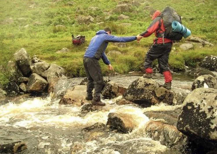 Two hikers crossing a stream in a rocky, grassy landscape, helping each other.
