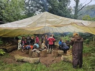 Group of people gathered outdoors under a large tarp in a forested area, some sitting on benches, others standing or walking, with backpacks and camping gear.