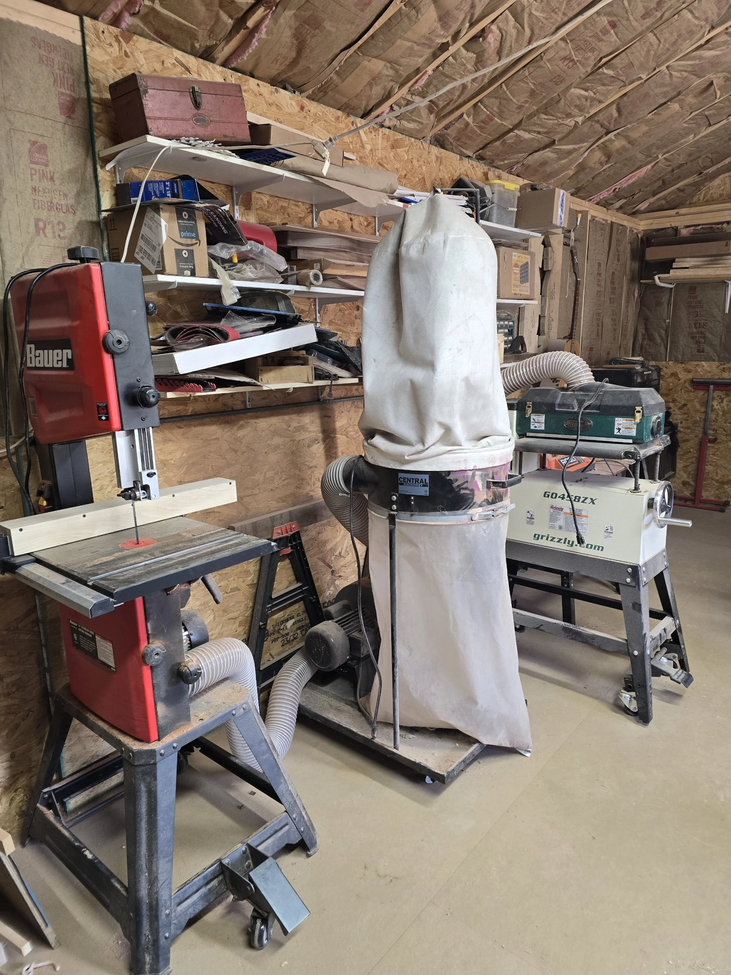 A woodworking workshop with two distinct woodworking machines connected to dust collection systems, shelves filled with tools and supplies, and a workstation with a saw.