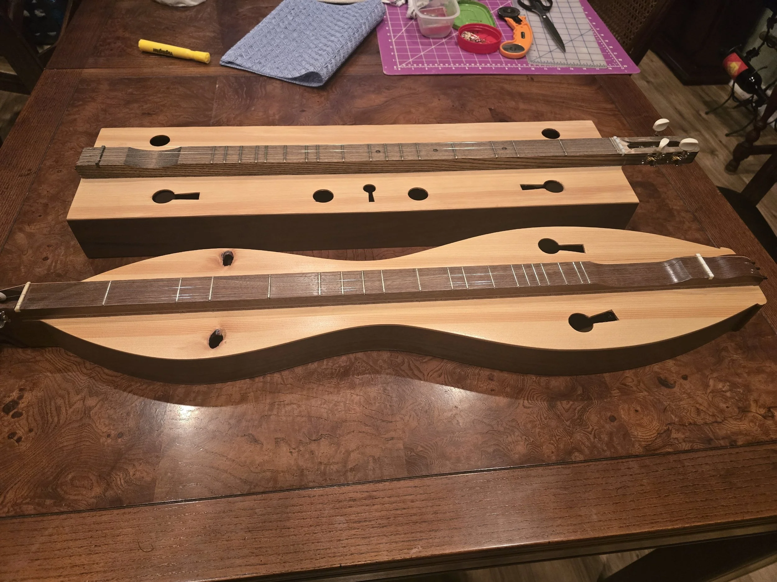 Two unfinished wooden guitar bodies on a table, with guitar necks attached, and various tools and supplies in the background.