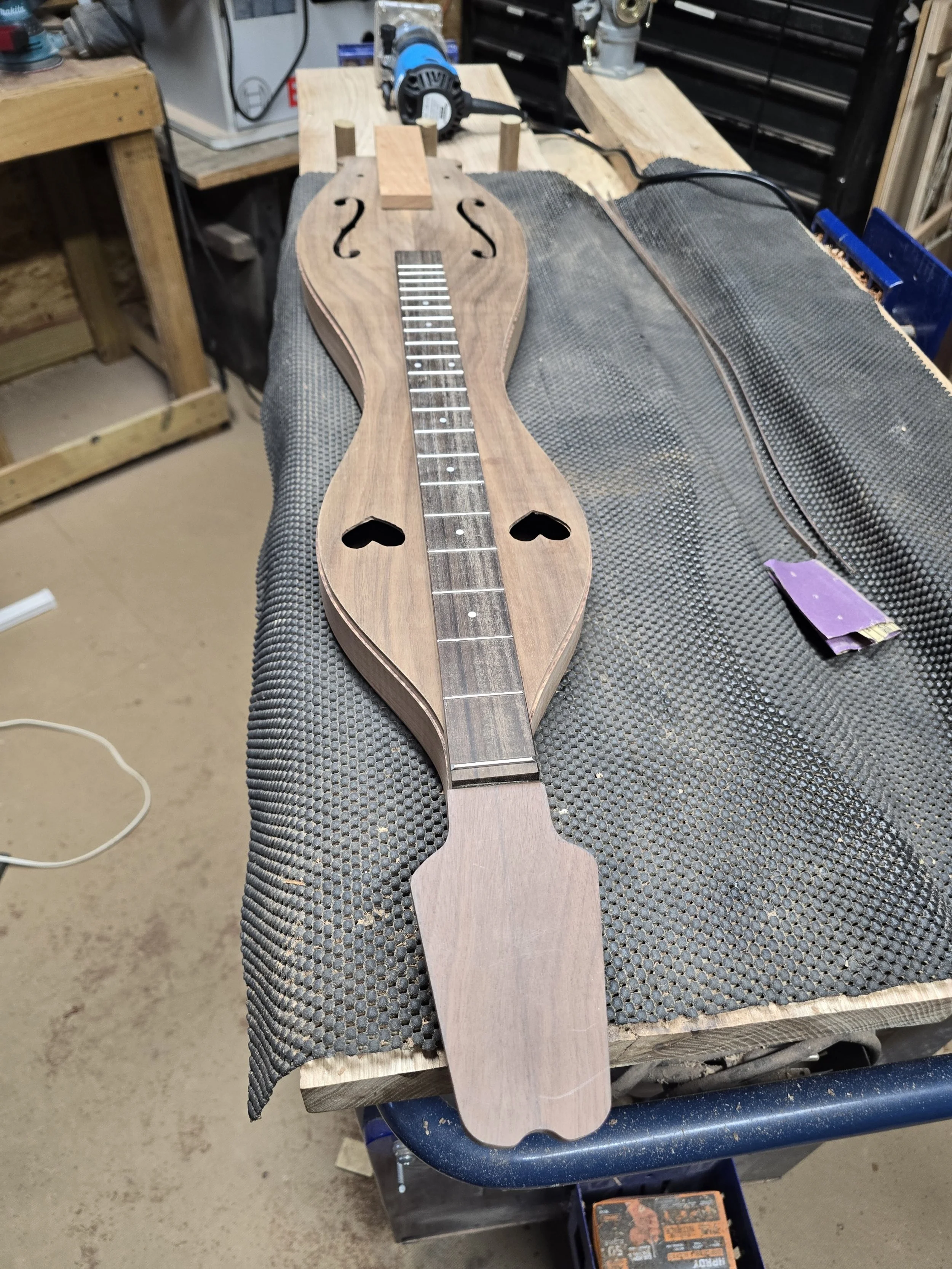 Unfinished wooden musical instrument resembling a ukulele or guitar, placed on a workbench in a woodworking workshop.