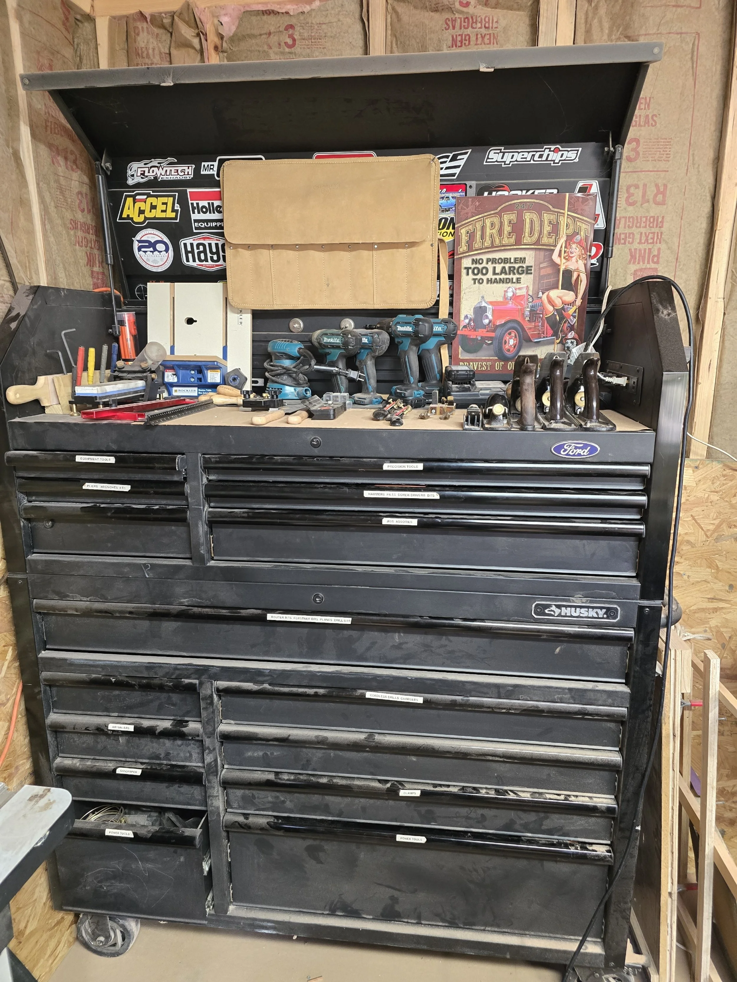 A black Husky tool chest with multiple drawers, positioned in a workshop with wooden walls. The top is cluttered with handheld power tools, a tan tool roll, a small white box, some markers, a red tool, and other small tools and parts. There are various stickers and signs in the background, including a vintage fire department poster and a stick-on label for organization.