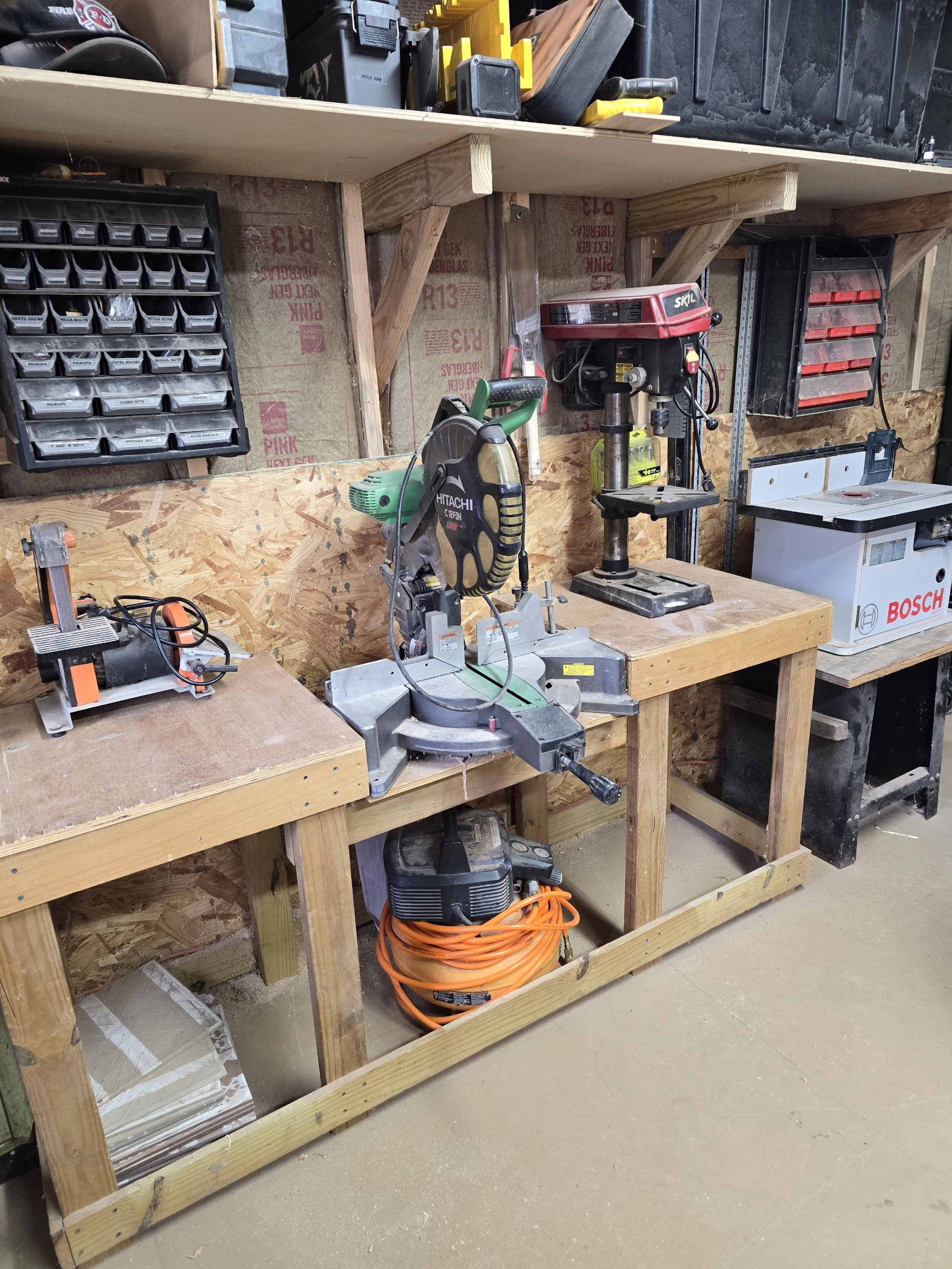 Workshop with various power tools on two wooden workbenches against a plywood wall, including a miter saw, drill press, and band saw, with storage bins on the wall and a coiled extension cord underneath.