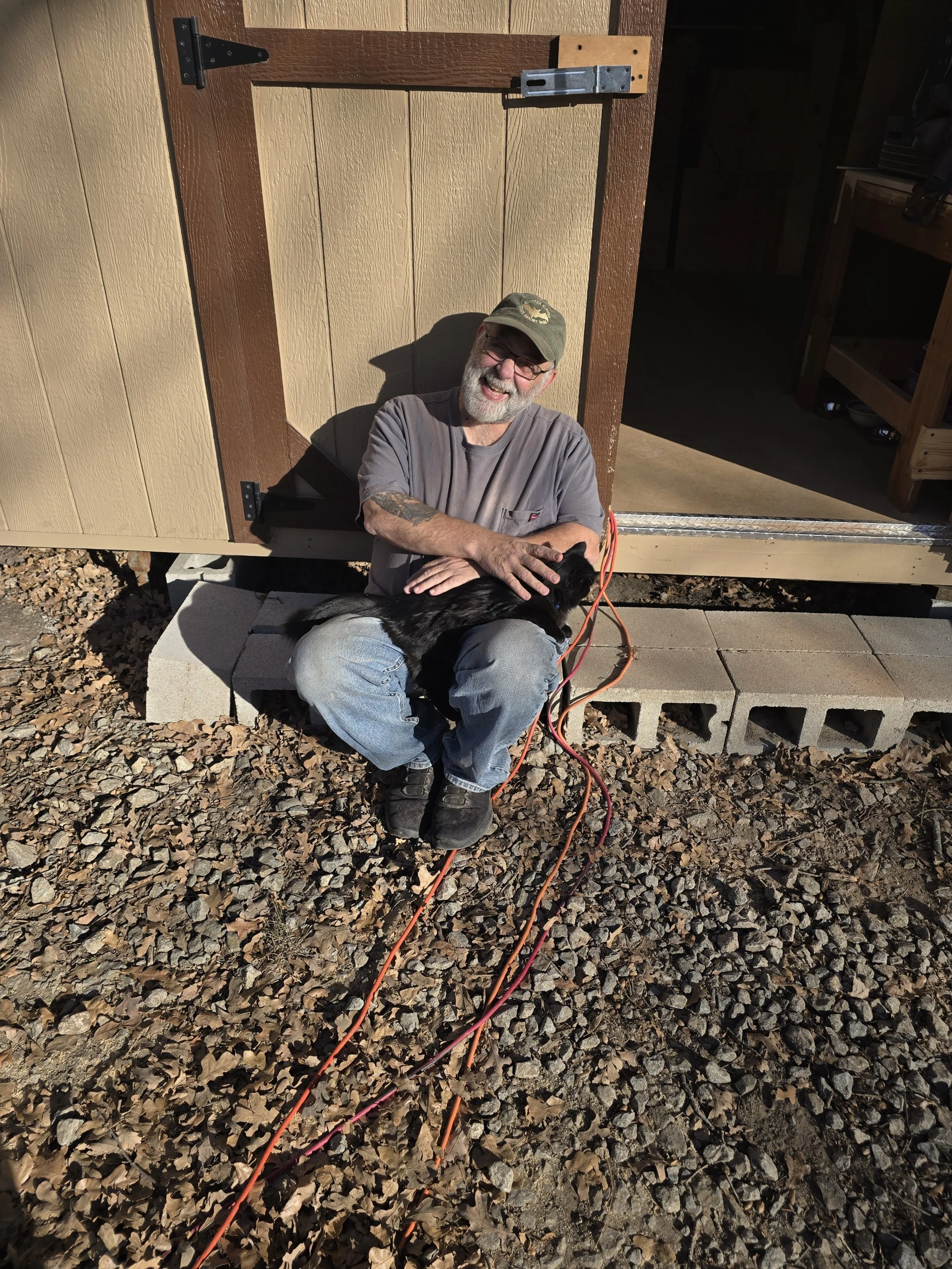 A man with a gray beard, glasses, and a baseball cap is sitting on a block of concrete in front of a small shed or building, smiling and petting a black dog.