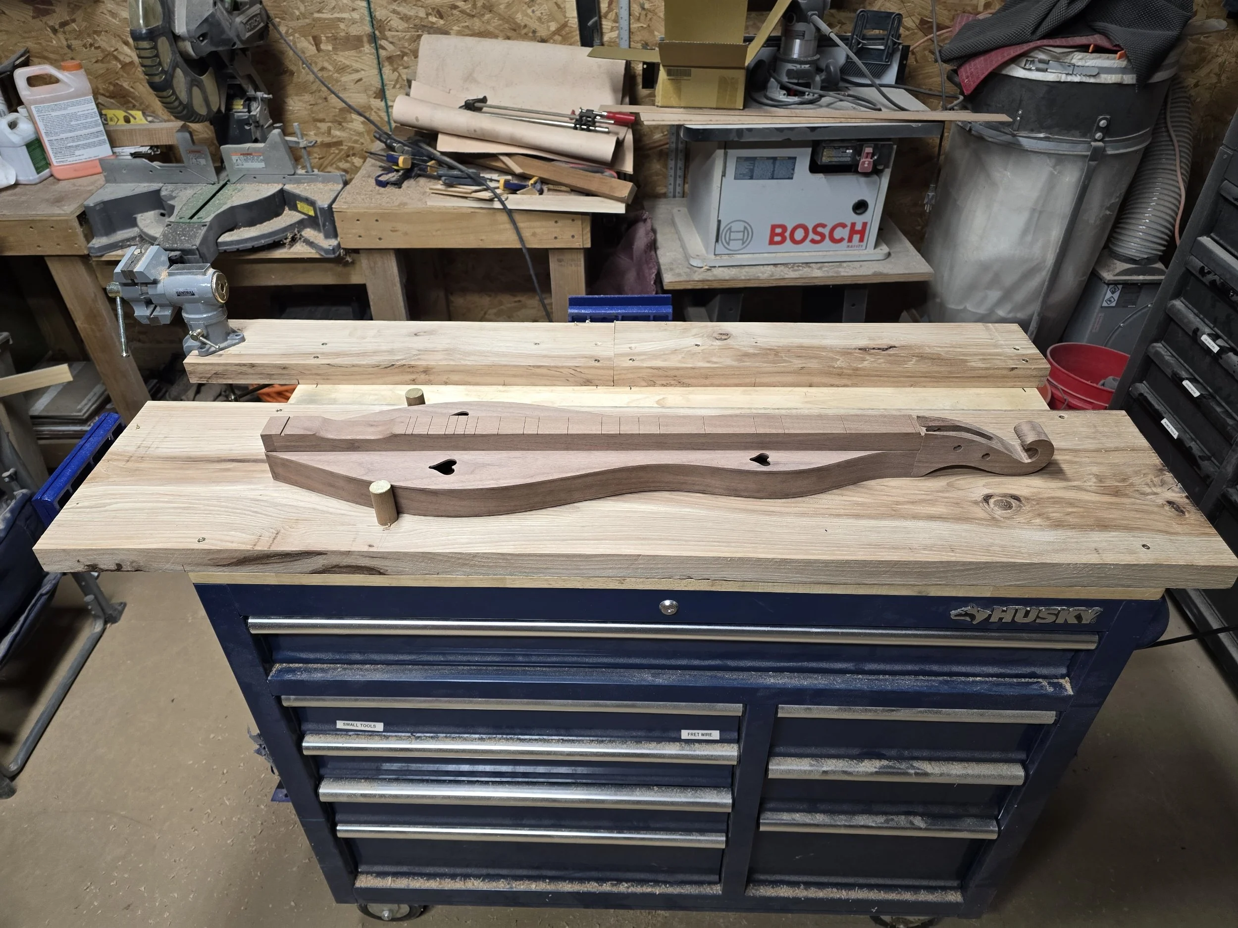 Wooden workbench with a carved wooden piece on top and woodworking tools in the background, including a Bosch tool and various supplies.