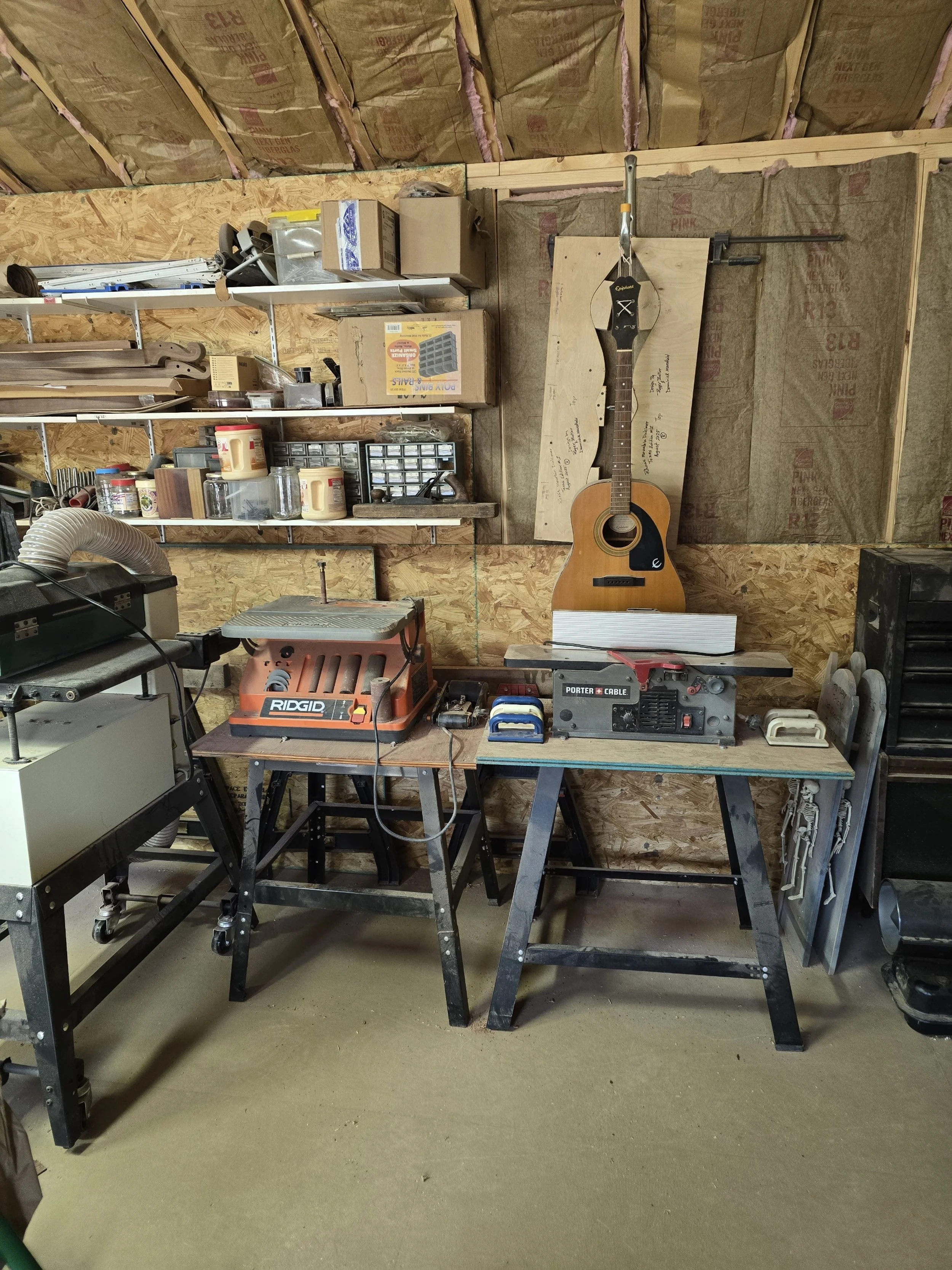 Workshop with tools and supplies including a guitar hanging on the wall, workbenches with a router and a planer, and shelves filled with jars, boxes, and small parts.