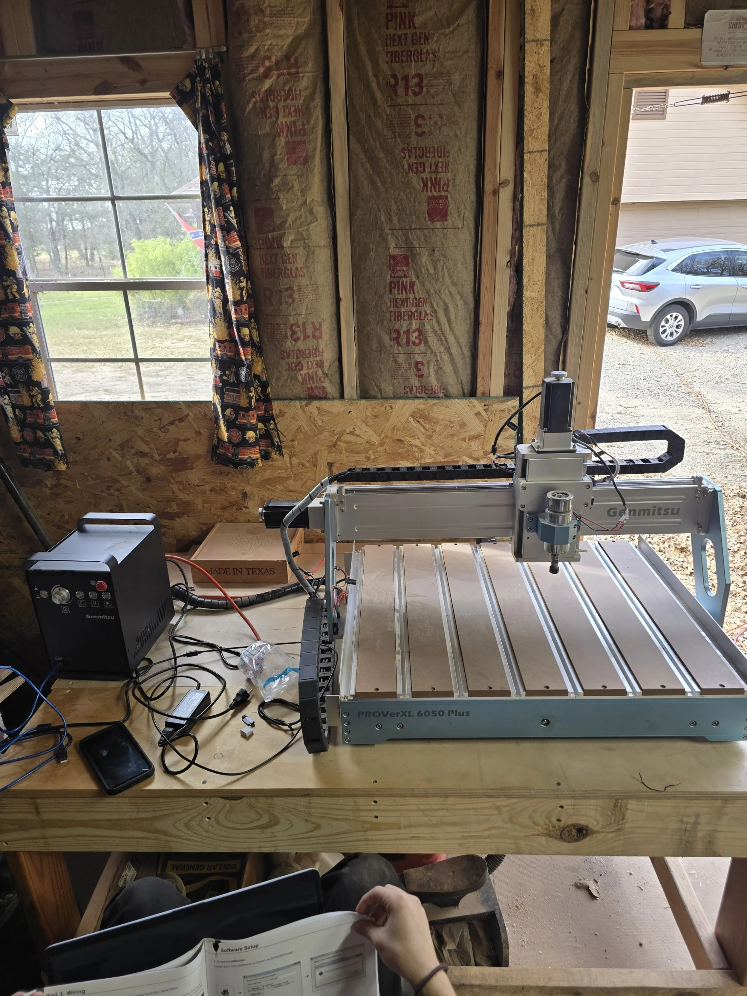 A CNC machine setup on a wooden workbench inside a garage or workshop. The machine is a Gennitsu PROverXL 6050 Plus model. There are various wires, a control box, and a plastic water bottle on the table. A person is seated at the table, holding a manual or instruction sheet.