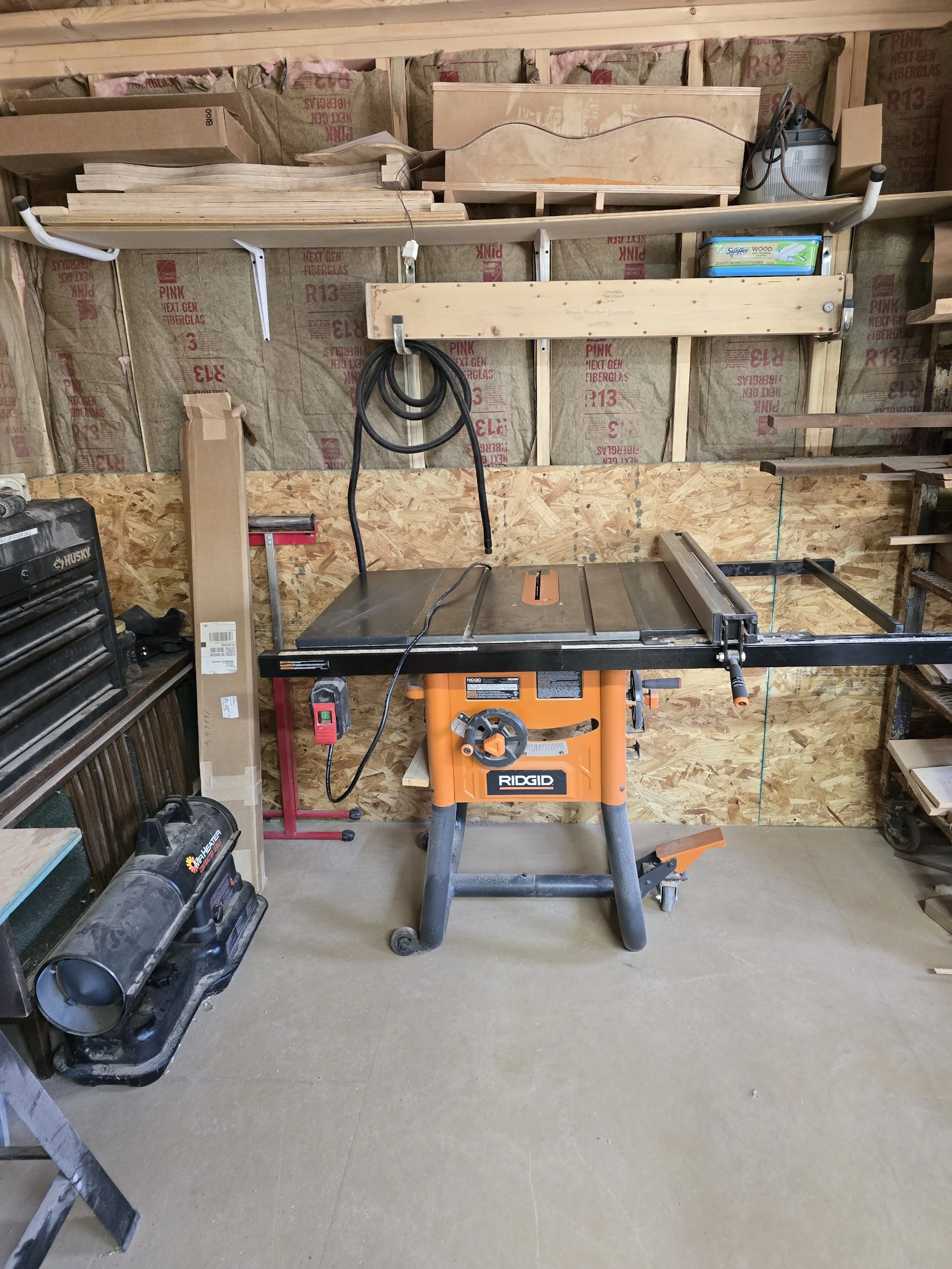 A woodworking workshop with a table saw, workbench, storage shelves, and various tools and supplies in a garage setting.