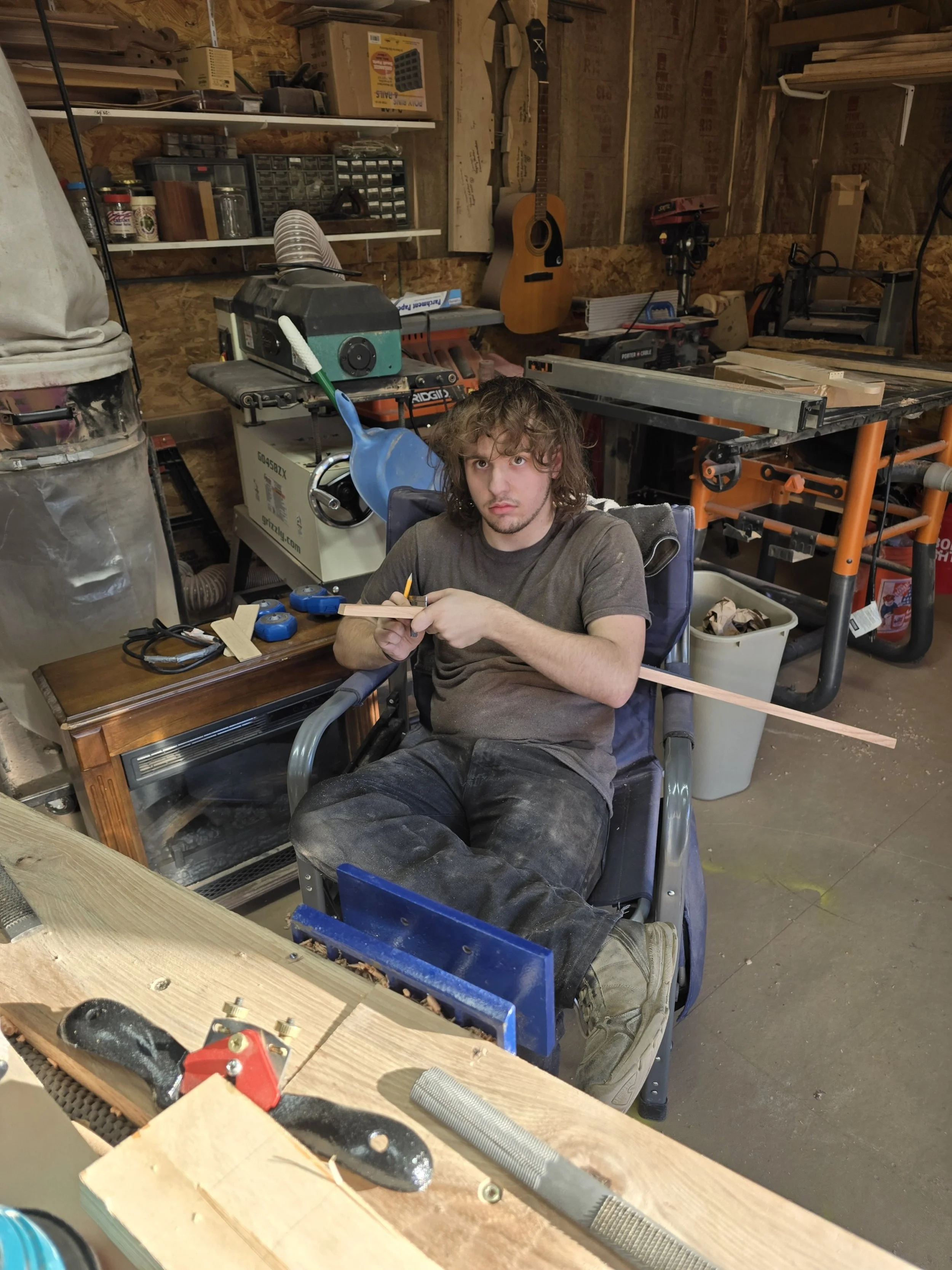 Young man in workshop sitting in wheelchair, working on a woodworking project with a wooden piece attached to his arm.