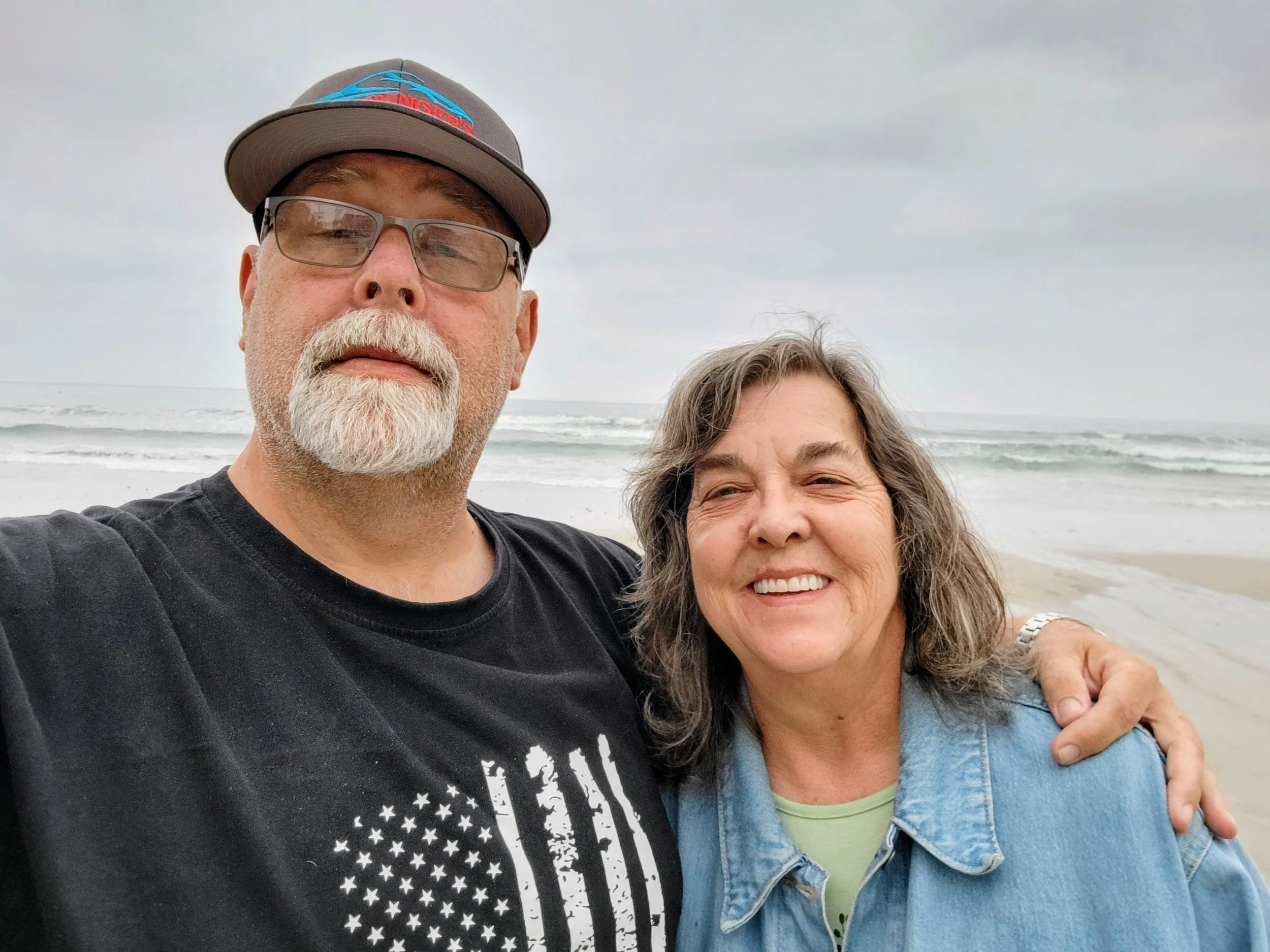 A man with glasses and a baseball cap taking a selfie with a woman at the beach on a cloudy day, ocean waves in the background.