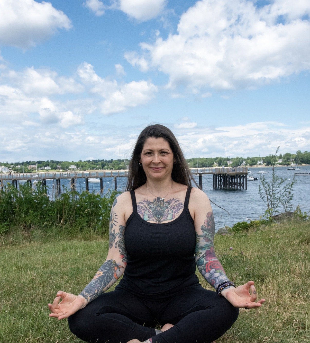 Woman with tattoos practicing yoga outdoors near a body of water under a partly cloudy sky.