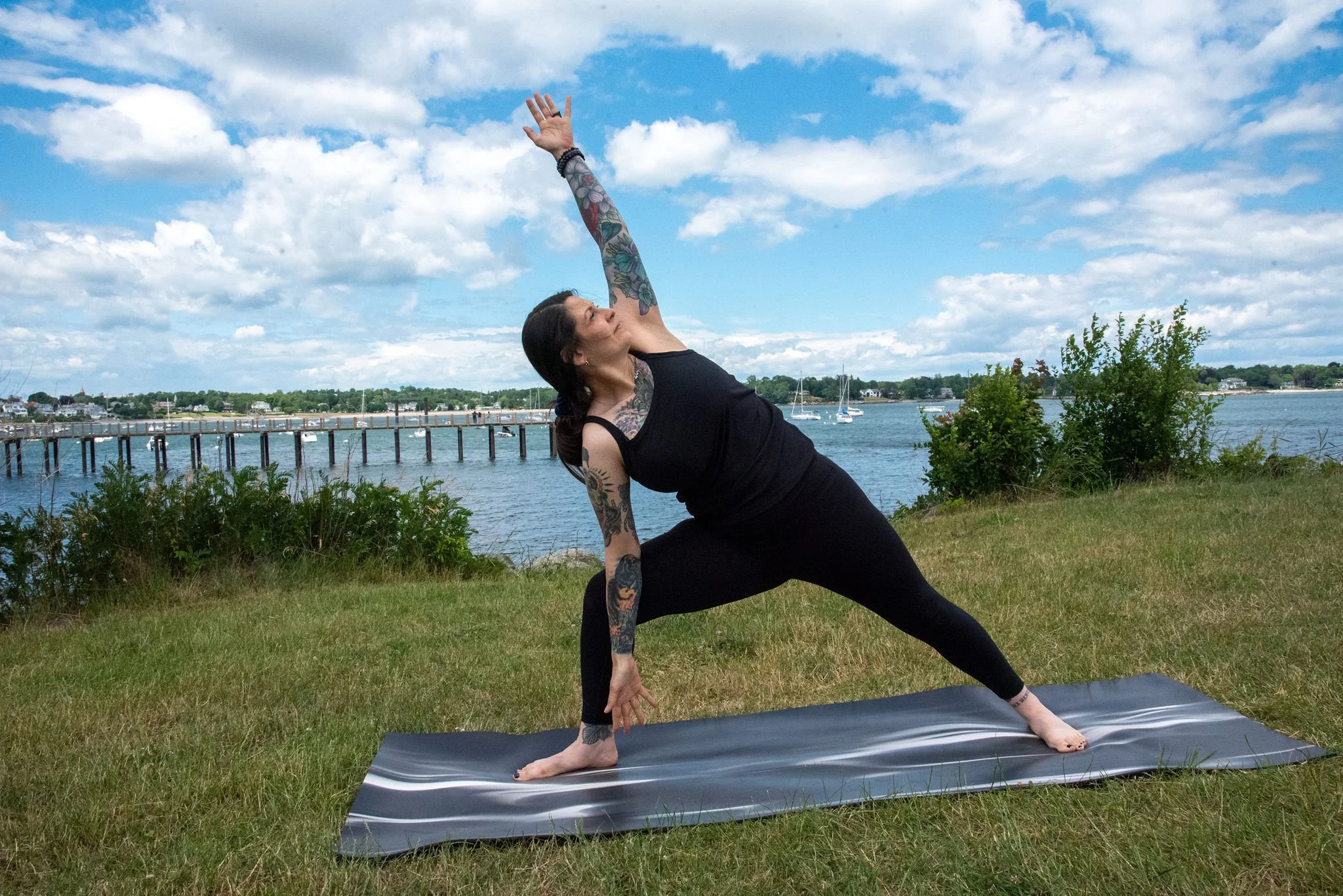 Woman practicing yoga outdoors on a mat near a river, performing a side stretch pose with her right arm extended overhead, wearing a black tank top and leggings, with tattoos on her arms and a scenic background of water, boats, and a blue sky with scattered clouds.