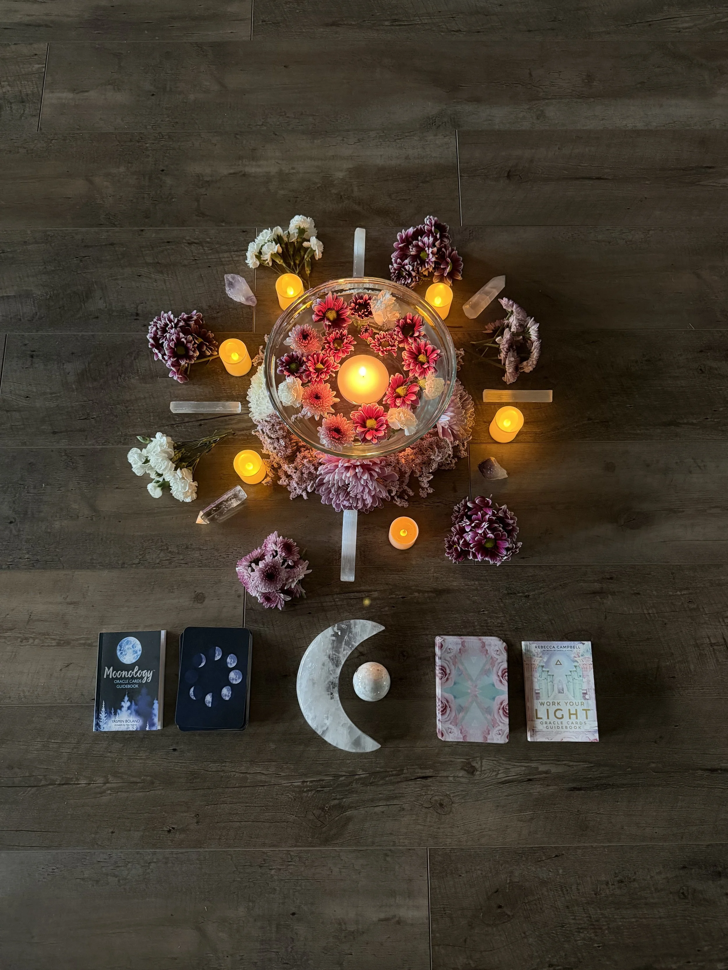 Flat lay of tarot and oracle cards, candles, flowers, and crystals on a wooden surface.