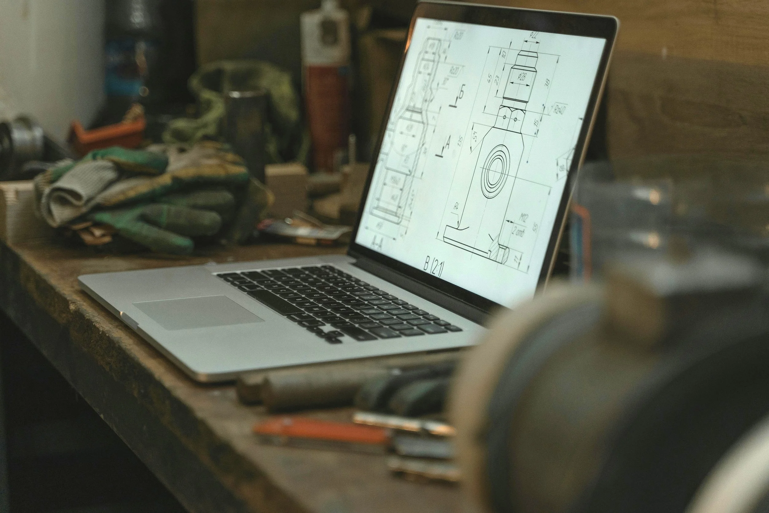 A laptop on a workbench displaying a technical engineering drawing or blueprint.