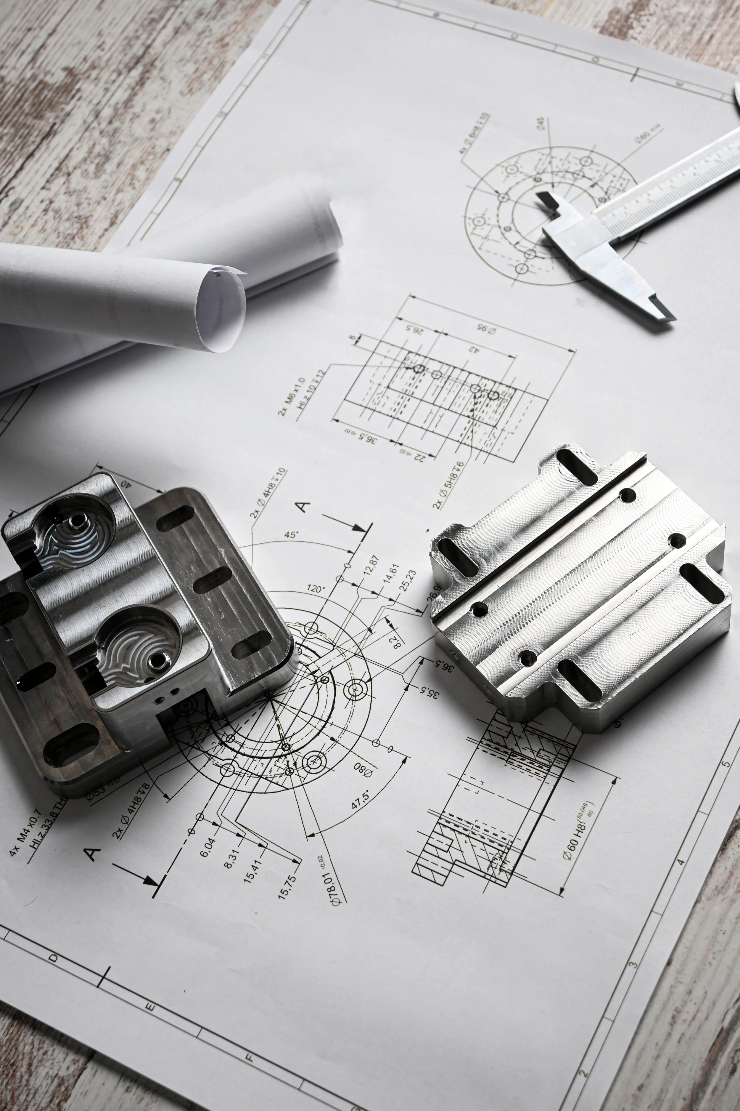 Technical engineering drawings along with metal machined parts, measuring tools, and a pencil on a wooden surface.