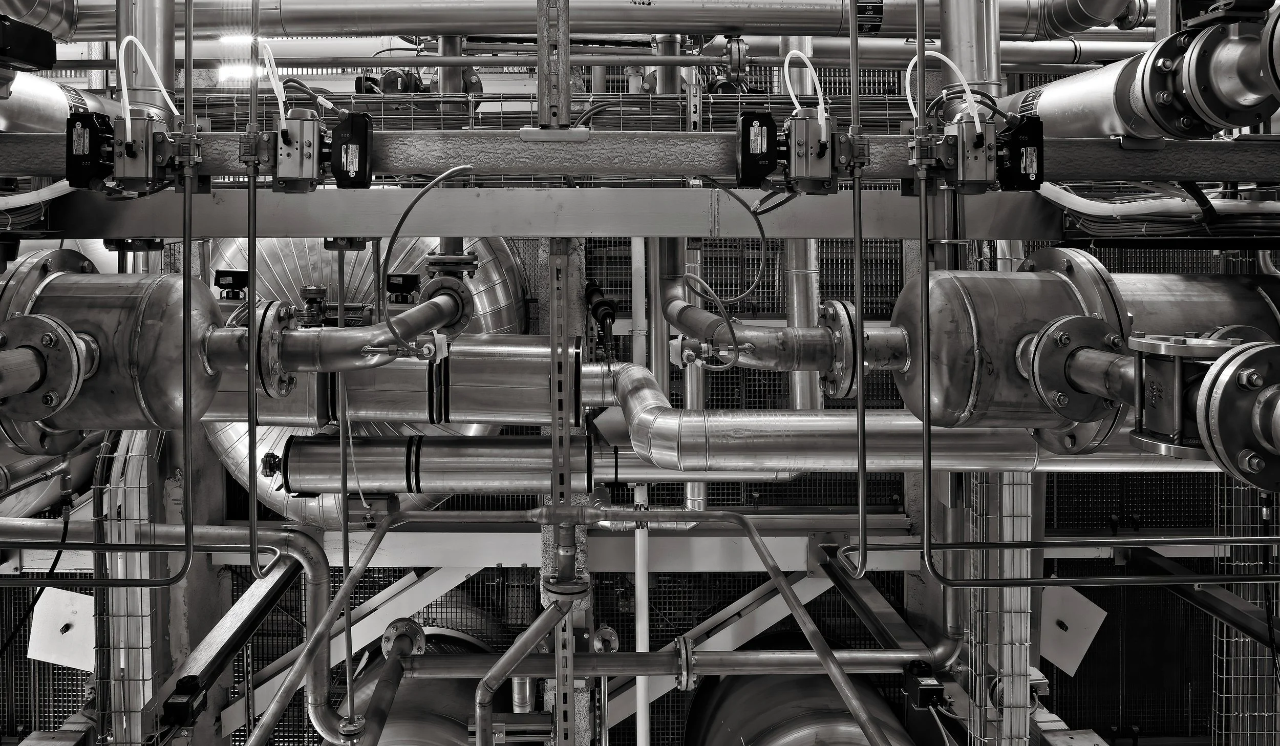 Industrial machinery with interconnected pipes and valves in a factory setting, black and white photo.