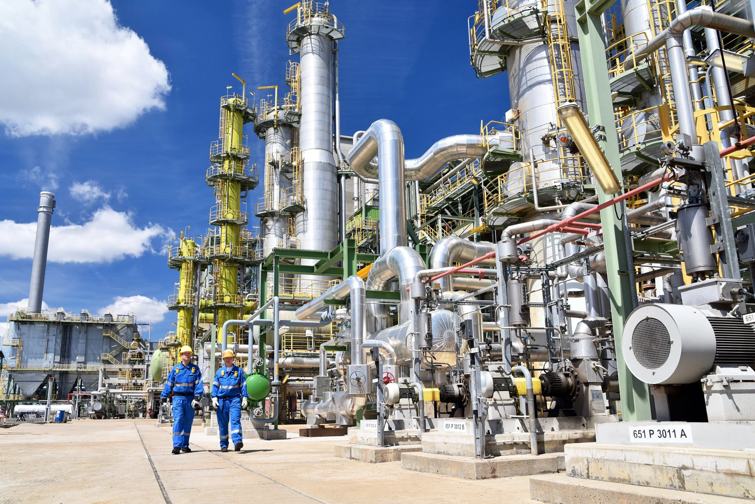 Two workers in blue uniforms and yellow helmets walking near industrial machinery in an oil refinery plant on a sunny day.