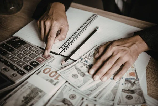 Person using a calculator with cash, a notepad, and a pen on a wooden table.
