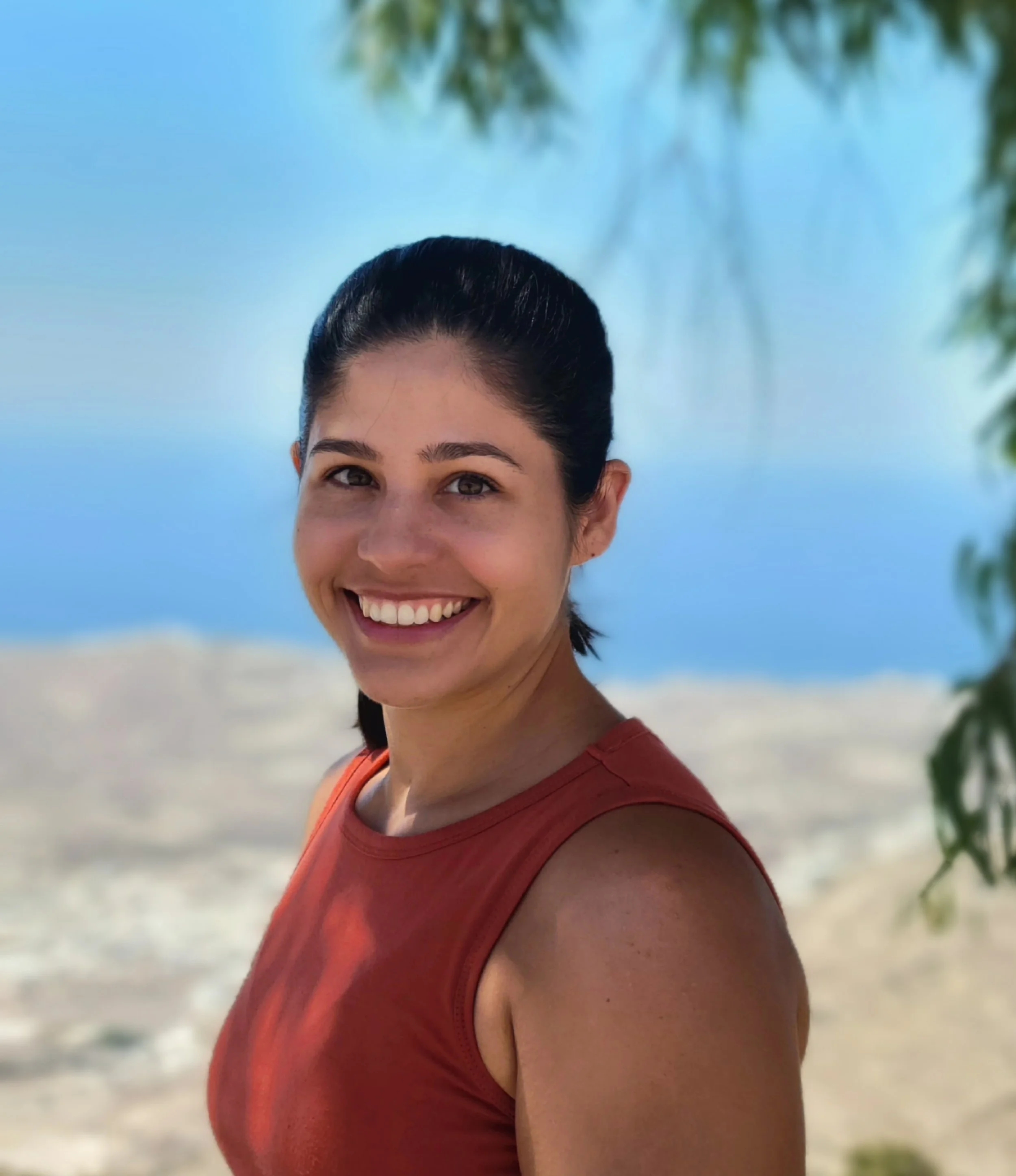 A person smiling on a beach, with part of their face and upper body visible, wearing a red sleeveless top, with a background of blue sky, blurred green foliage, and sandy shore.
