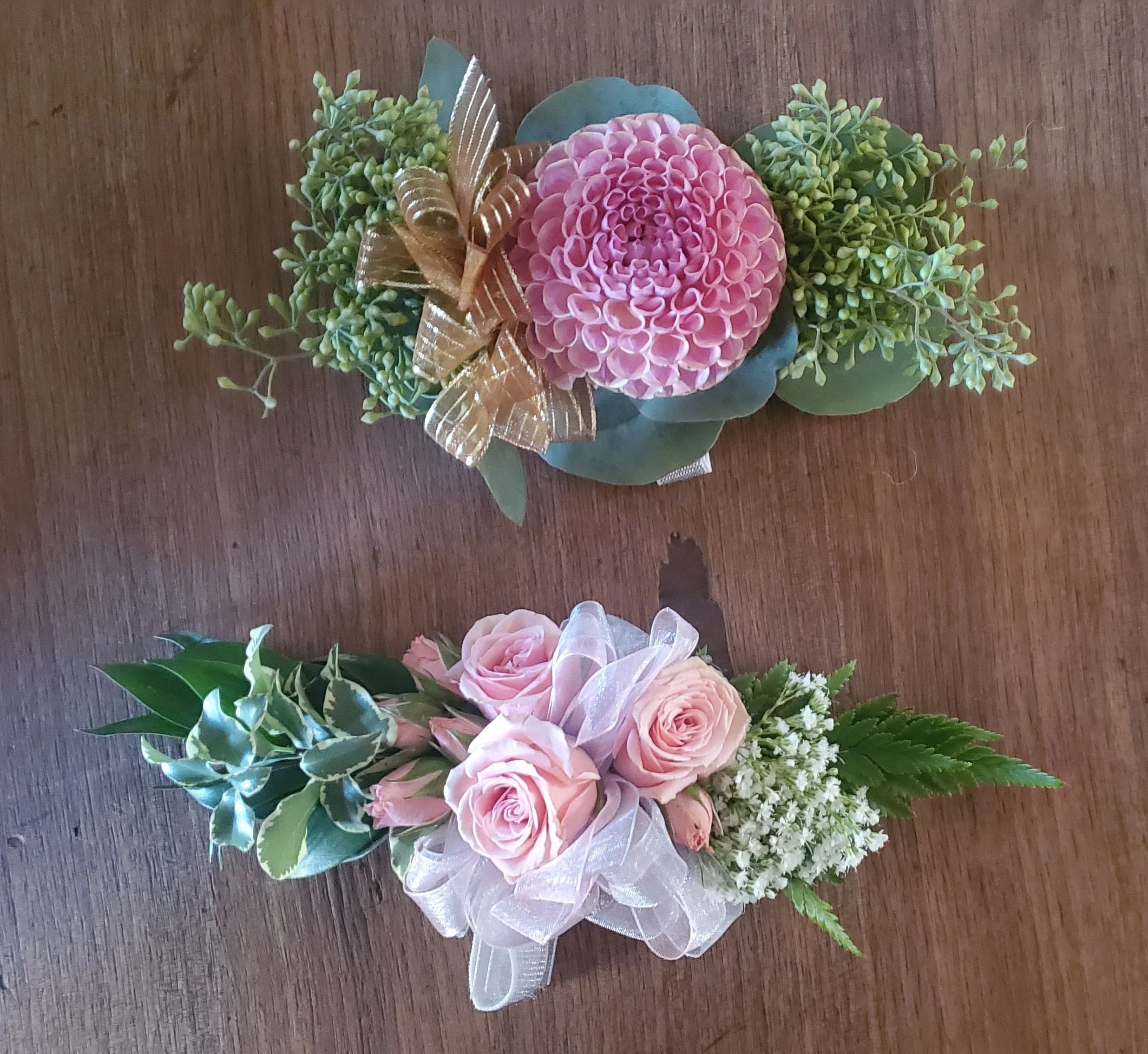 Two floral arrangements on a wooden surface, one with pink and green flowers and a gold bow, the other with light pink roses, white flowers, and green leaves.