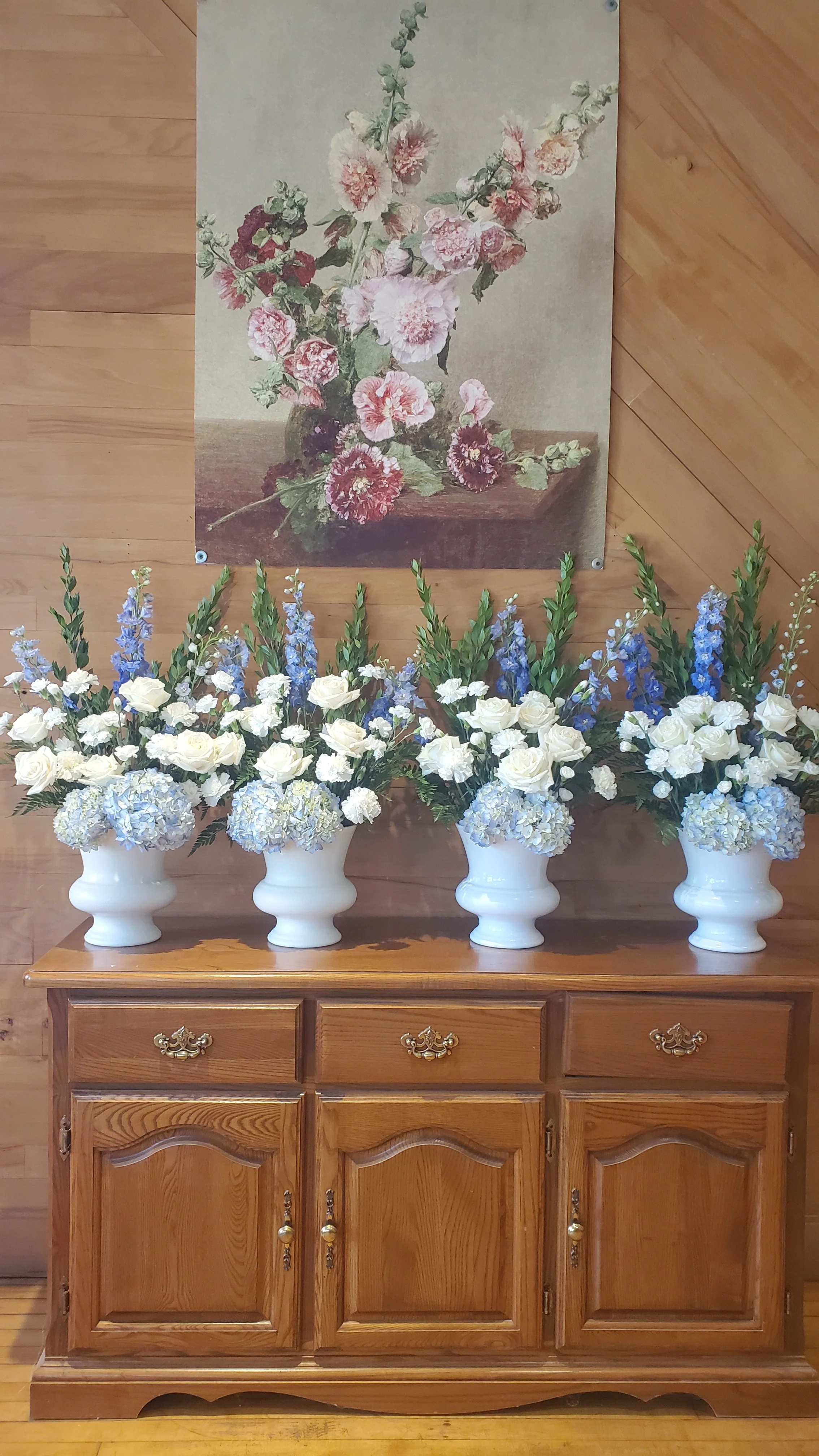 Four white vases filled with white roses and blue hydrangeas, and tall blue delphinium flowers, placed on a wooden sideboard against a wood-paneled wall. Above, a floral painting of pink and red carnations in a vase hangs on the wall.
