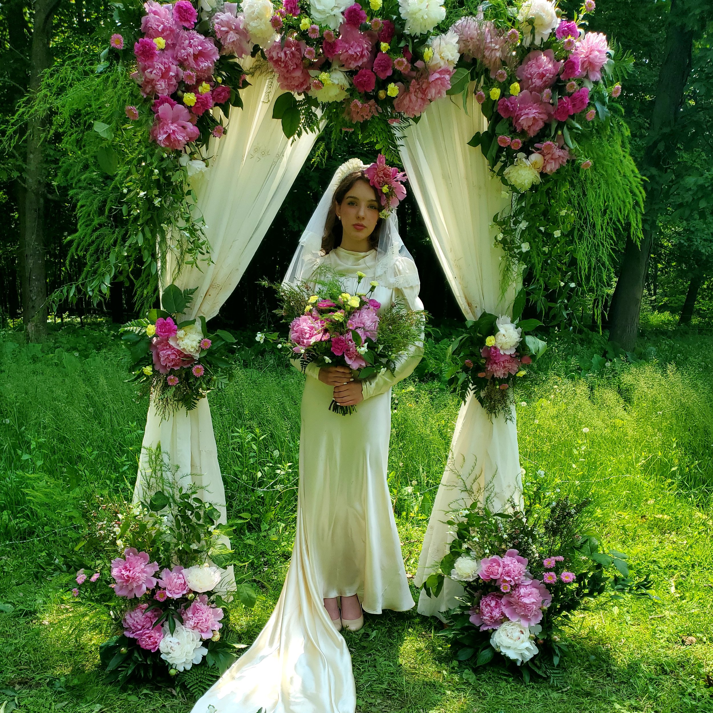 A bride standing outdoors under a floral arch, holding a bouquet, surrounded by greenery. The arch is decorated with pink, white, and purple flowers and white curtains.