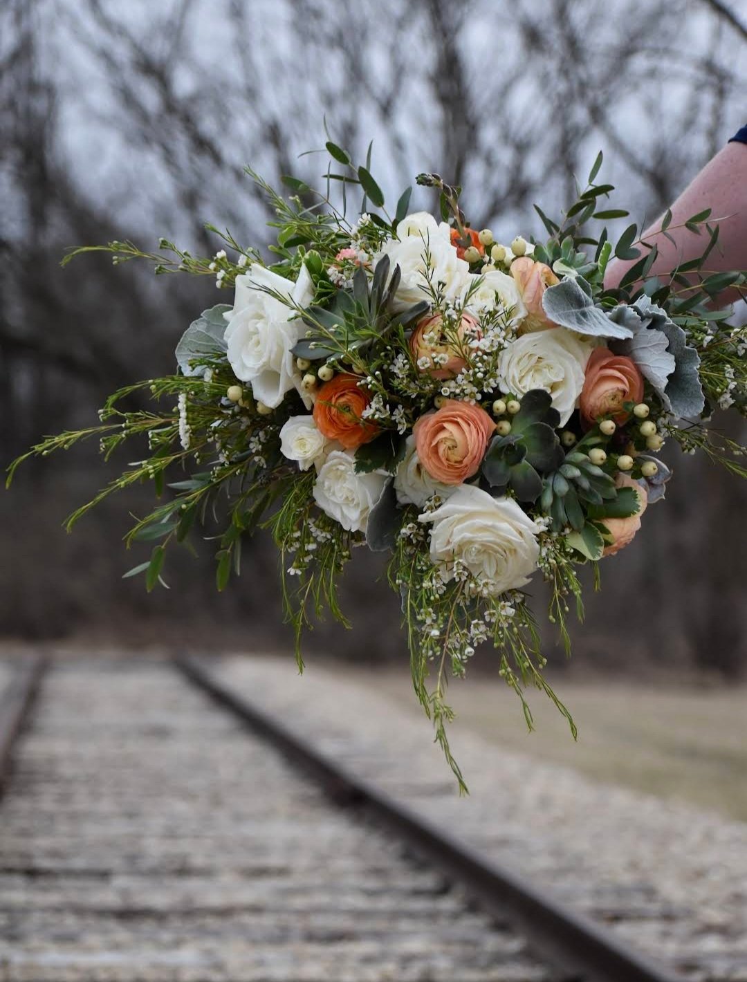 Person holding a bouquet of white and peach flowers outdoors with blurred trees and railroad tracks in background.