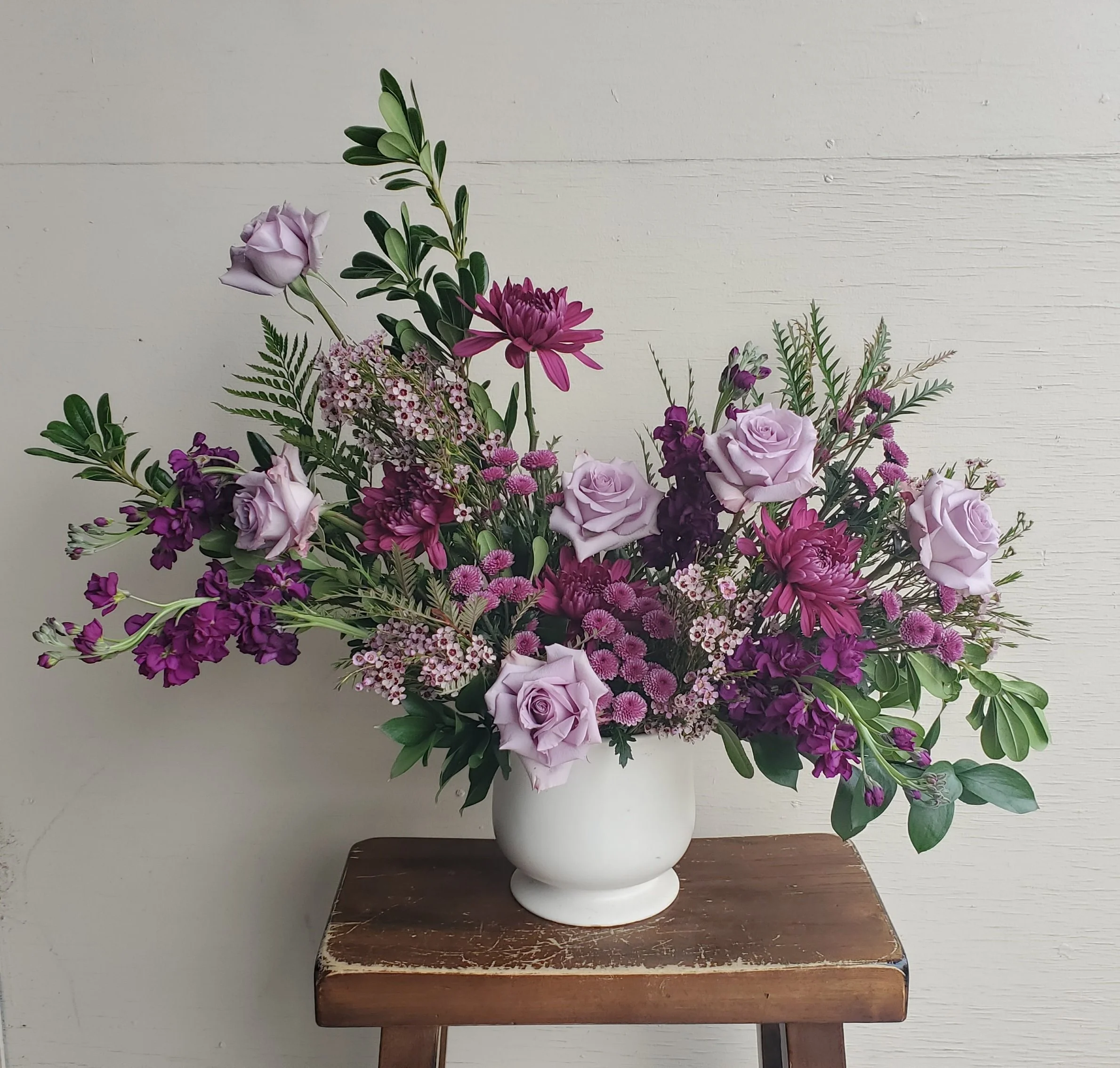 A floral arrangement with purple roses, pink daisies, and various greenery in a white vase on a wooden stool.