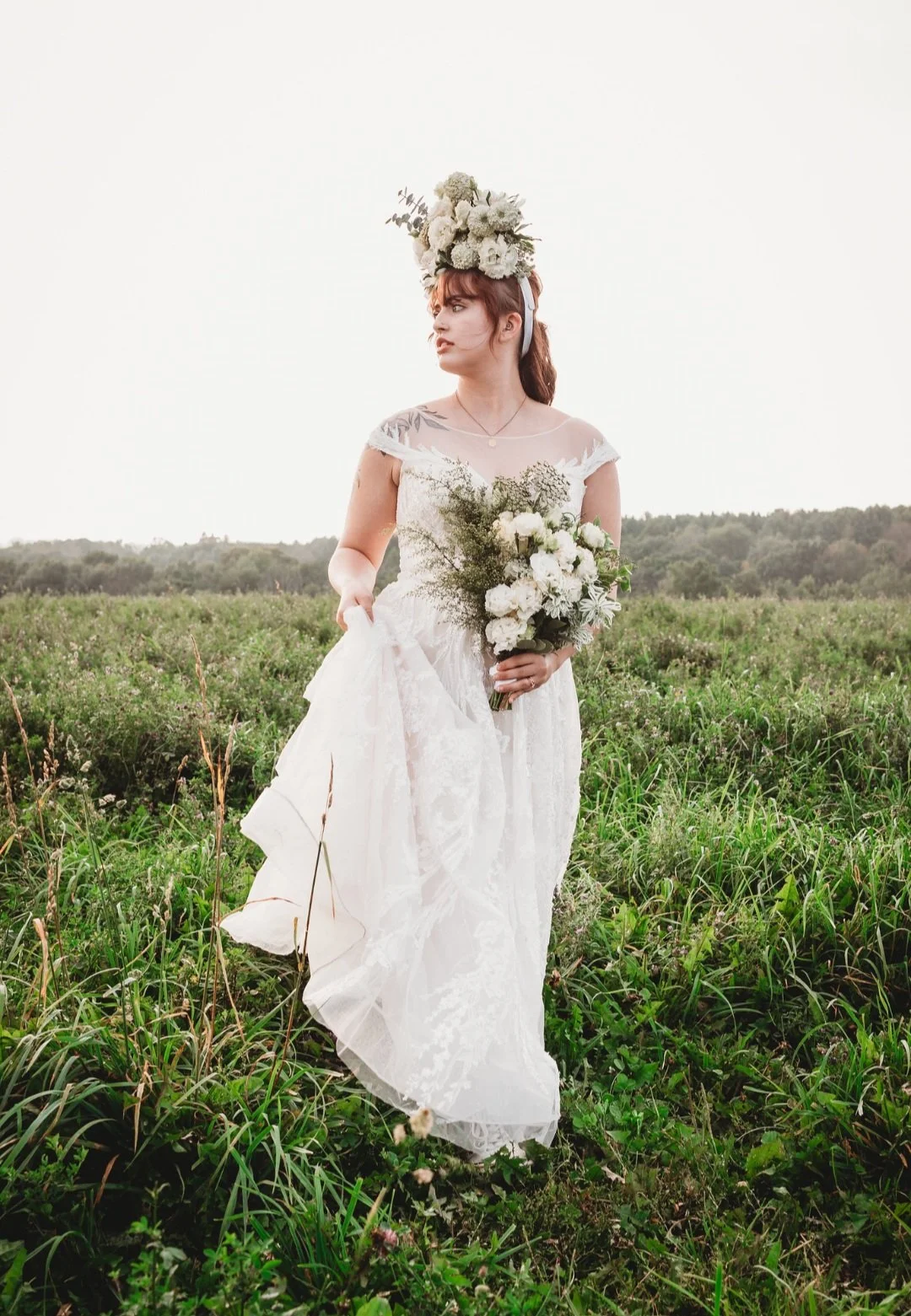 A woman in a white wedding dress holding a bouquet of white flowers and greenery, standing in a green field with a cloudy sky in the background.
