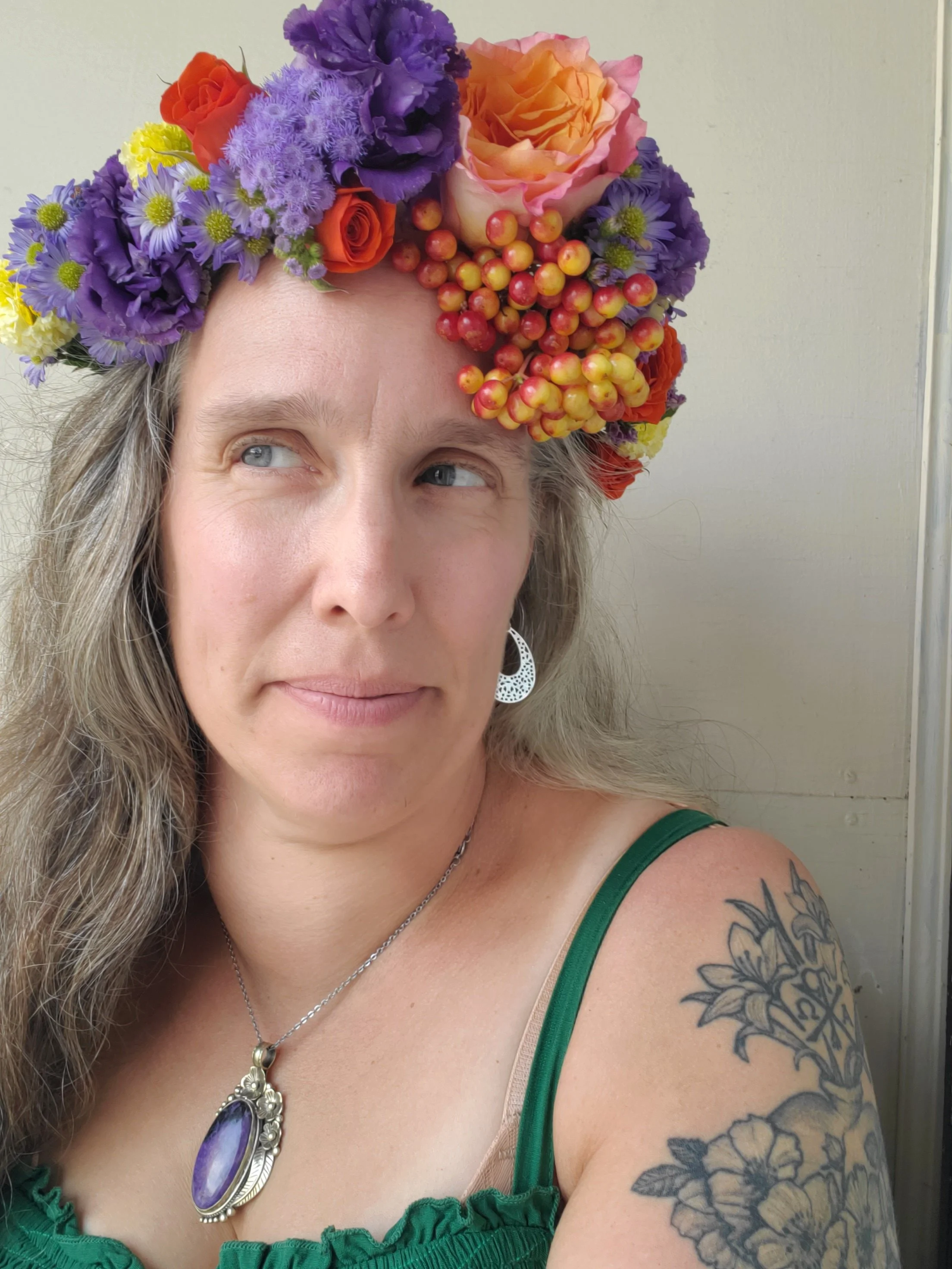 Woman with long blonde hair wearing a flower crown made of colorful flowers, a green top, and jewelry.