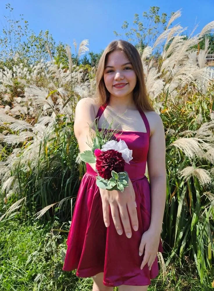 A young woman in a maroon dress stands outdoors in a field of tall, feathery grasses, smiling and showing a wrist corsage with maroon and white flowers and green leaves on her extended arm.