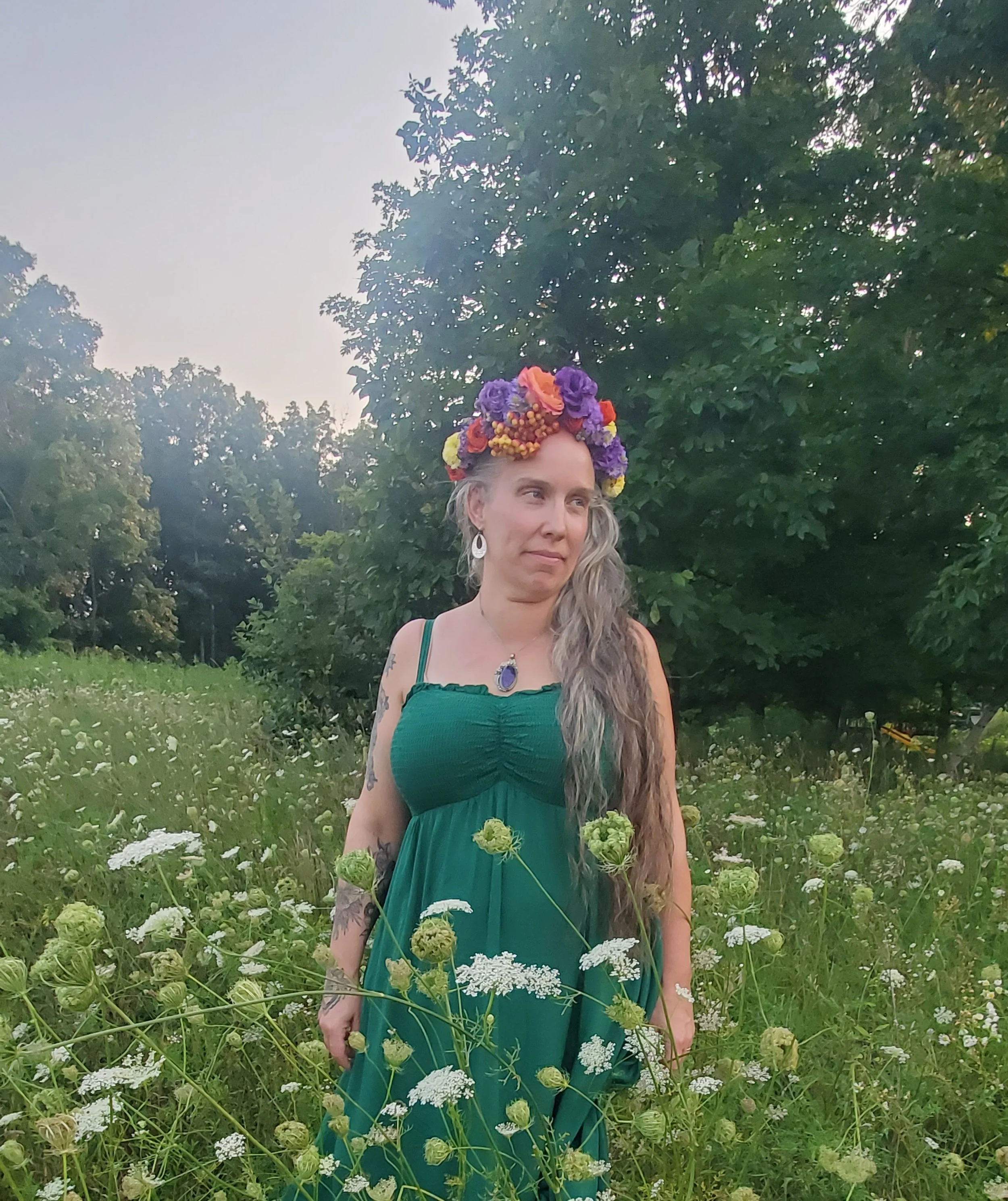 A woman with long, wavy gray hair stands in a field of white and green flowers, wearing a teal dress and a colorful floral crown, with a large purple pendant necklace, surrounded by green trees.