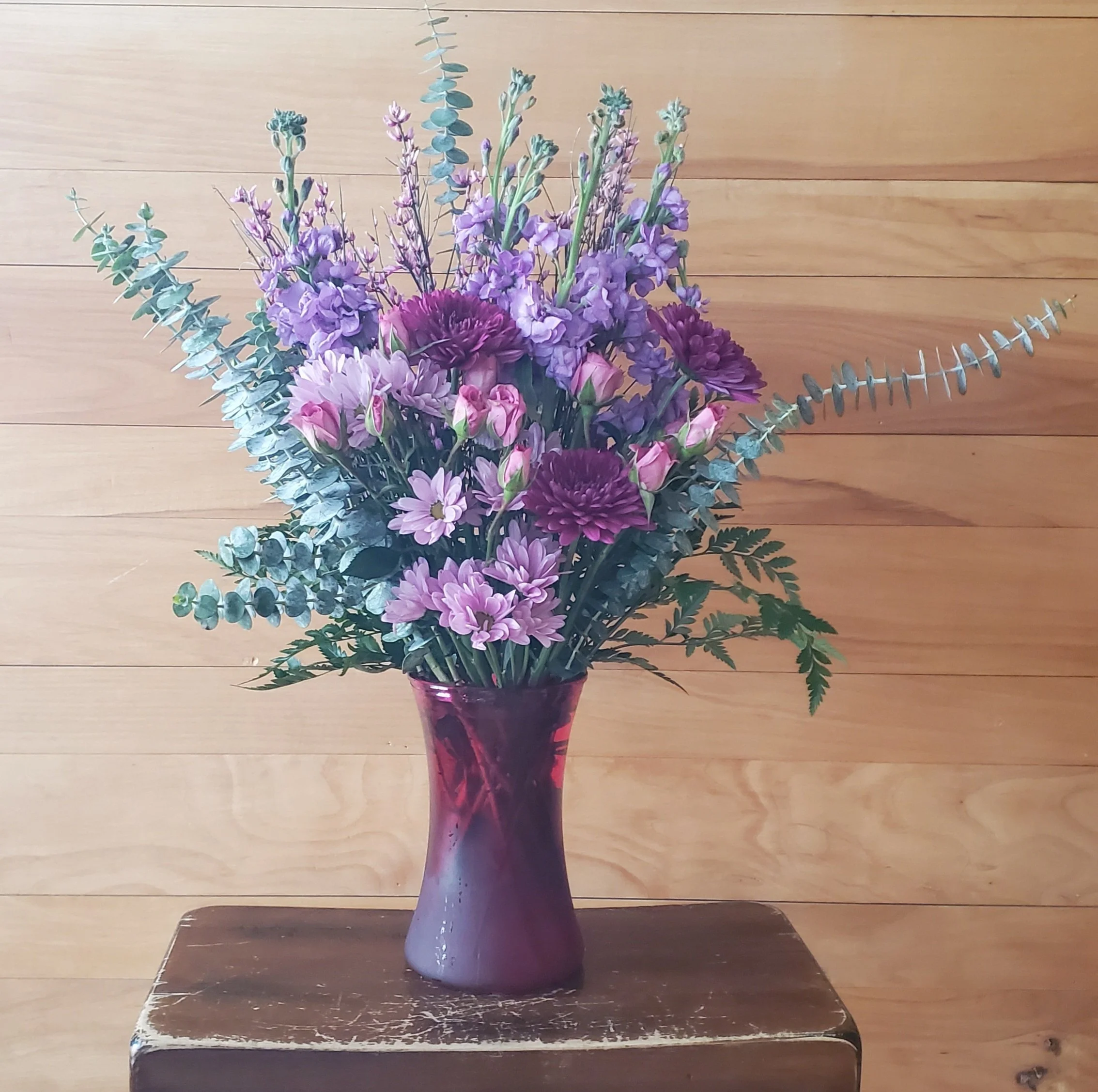 A bouquet of purple and pink flowers in a purple vase on a wooden table against a wooden wall background.