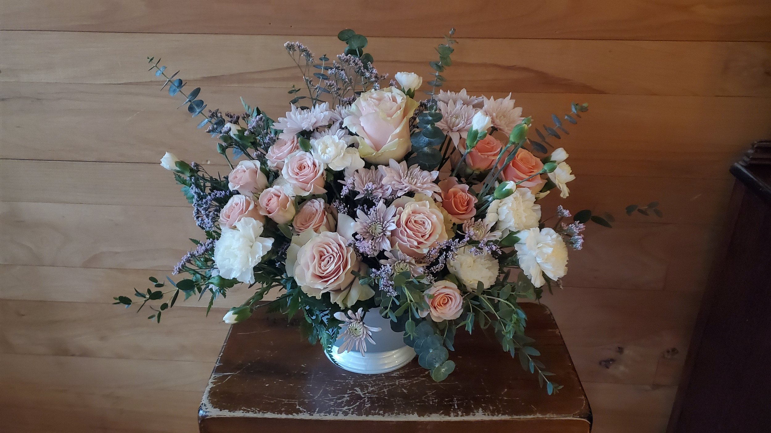 A large floral arrangement with peach, white, and light pink roses, white carnations, and purple accents in a white vase on a wooden table against a wood-paneled wall.