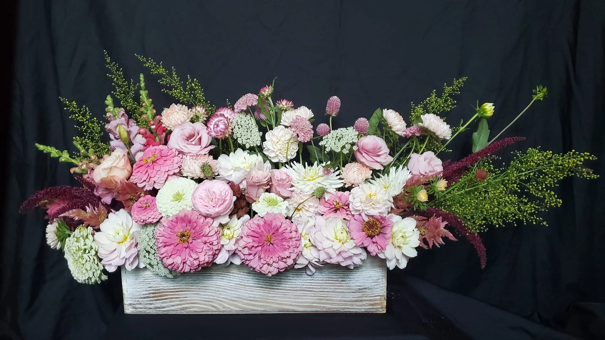 A floral arrangement with pink, white, and purple flowers in a rectangular wooden container against a black backdrop.