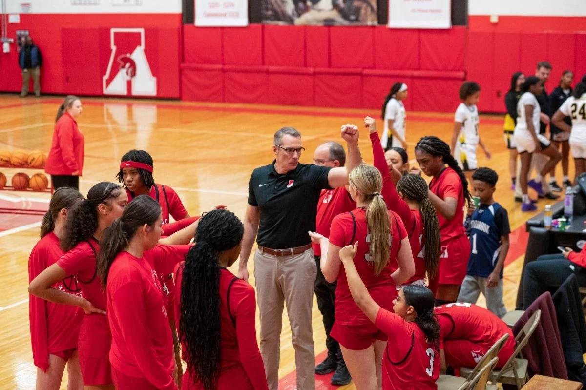A girls' basketball team in red uniforms huddles around their coach, Tim Corrigan, during a timeout in a gymnasium.